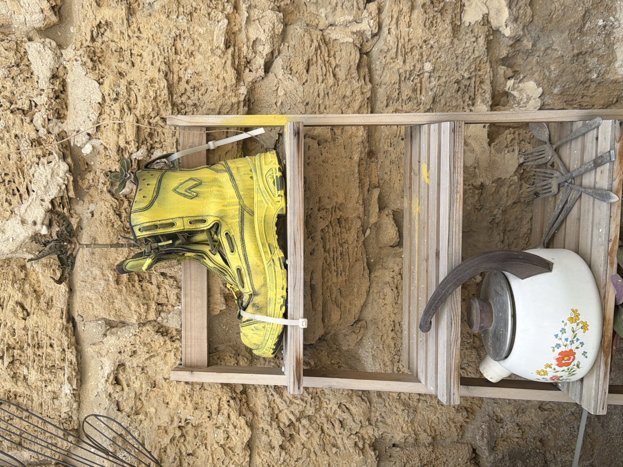 A vertical shot captures a rustic, charming display of repurposed household items against a heavily textured, light brown stone or concrete wall. The wall, possibly an aged building facade, shows significant erosion and numerous small holes, indicating long-term outdoor exposure.
Central to the image is a simple, light-colored wooden frame, resembling a small ladder or a deconstructed shelf unit, mounted against the wall. On the left side of this frame, a bright yellow boot or heavy-duty shoe is prominently displayed, secured by a white zip tie. The boot features black outlines and a stylized, chevron-like symbol on its side.
To the right, a white enamel kettle with a dark, curved handle and a metal lid is positioned within the wooden frame. The kettle is adorned with a cheerful floral pattern in red, orange, yellow, and blue with green leaves. Above it, several aged metal utensils, including forks and spoons, are fastened to the wooden slats, appearing tarnished with time.
Further details include thin, dried-out tendrils or wires extending across the upper left of the wall, and a small succulent plant growing near the yellow boot. Part of a metal whisk is visible in the bottom-left corner. The clear, natural lighting suggests it is daytime. The arrangement evokes a sense of resourceful decoration or an artistic installation, typical of the creative street aesthetics found in cities like Tel-Aviv. No discernible text is visible.