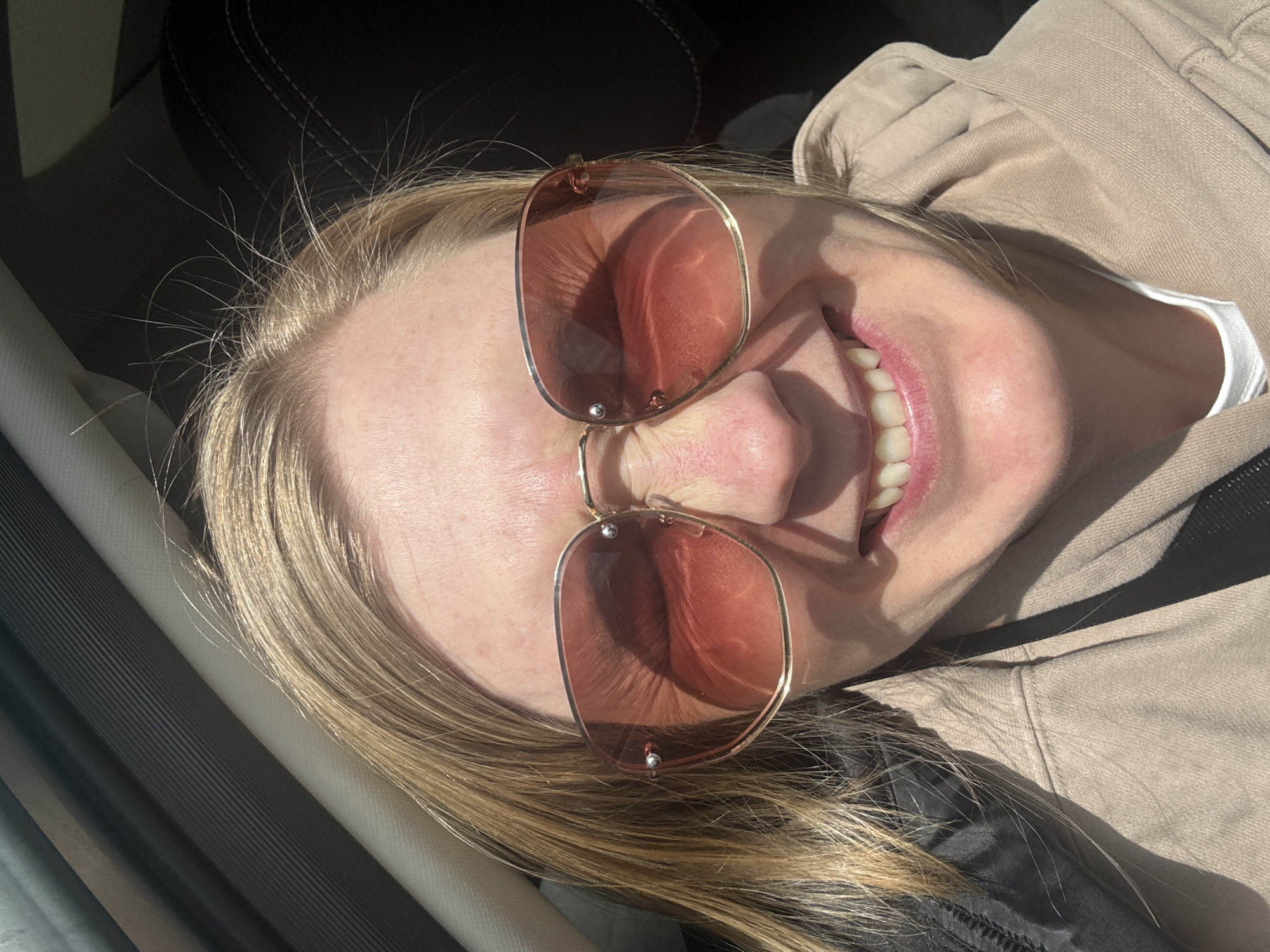 A Caucasian woman with blonde hair and fair skin is smiling while wearing rose-tinted sunglasses. She is wearing a beige hooded sweatshirt and possibly seated in a car. The exact location within Mar'evka, Russia, cannot be determined from the provided image.