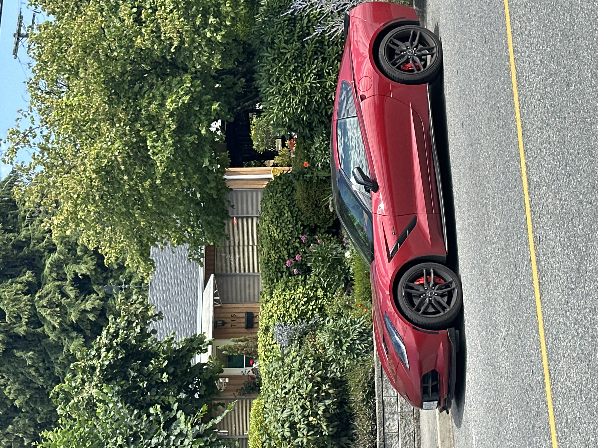 A cherry red Chevrolet Corvette is parked on the side of a residential street in Coquitlam, Canada. The car is positioned parallel to the curb, facing right. The front of the car is slightly angled towards the viewer, revealing its sleek headlights and aerodynamic design. The vehicle has black alloy wheels with red brake calipers visible behind the spokes.
Behind the car, a well-maintained yard is visible. A low, grey stone wall separates the street from the yard. A dense hedge runs along the top of the wall, and beyond that, a variety of plants and flowers bloom in shades of pink and purple. Further back, a single-story house with wooden siding and horizontal blinds on the windows is partially visible. The house number "715" is displayed near the entrance. Lush green trees provide a backdrop to the scene, with some branches extending over the house and yard. The sky is a clear blue, suggesting a sunny day. A yellow line painted on the asphalt road indicates a traffic lane.