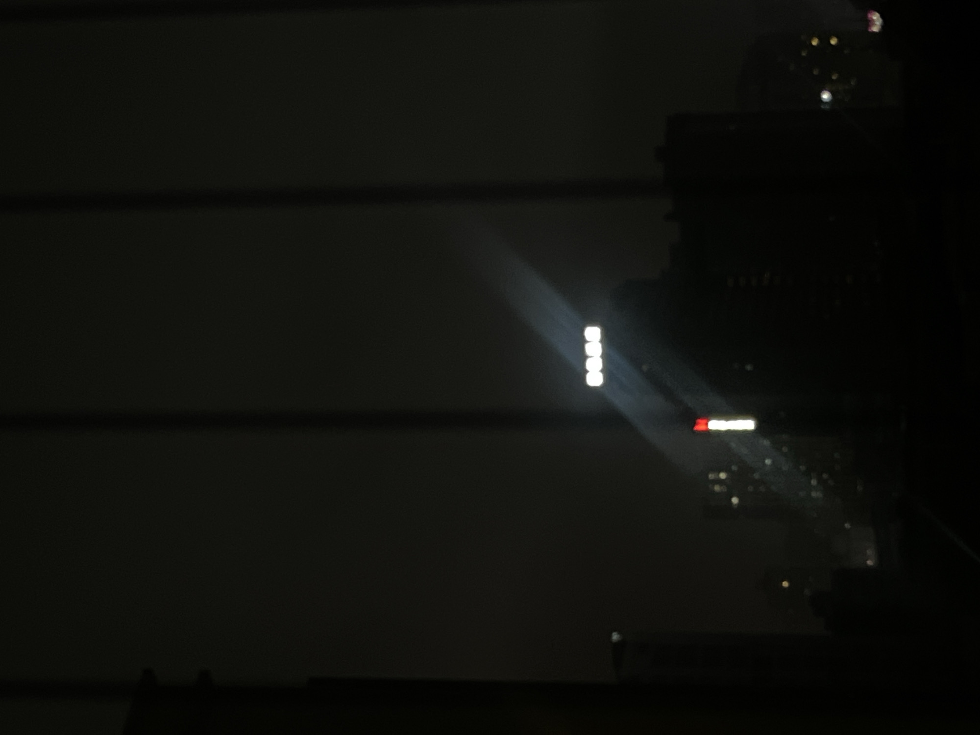 The image displays a nighttime urban landscape in Jiulongpo District, China, viewed from an interior perspective through what appears to be a window. Two dark, vertical bands, consistent with window mullions or blinds, divide the frame, appearing approximately one-third and two-thirds across from the left edge. The scene is dominated by darkness, with artificial light sources providing the primary illumination.
In the middle-ground, a bright, horizontally oriented illuminated sign displays the English word "DOES" in white characters. This sign is positioned slightly above the horizontal center of the frame and serves as a significant light source. Below and to the left of the "DOES" sign, a vertically oriented light fixture is visible, featuring a red rectangular segment atop a longer white rectangular segment.
Distinct rays of light emanate diagonally upwards and to the right from the "DOES" sign, suggesting light scattering through atmospheric particles or lens flare. Scattered, blurred points of light (bokeh) are visible in the background, particularly towards the lower right and left, indicating distant city lights. Silhouettes of indistinct buildings are visible against the uniformly dark sky. No individuals, specific actions, or interactions are discernible within the frame. The environment is an urban night scene under very low ambient light conditions.