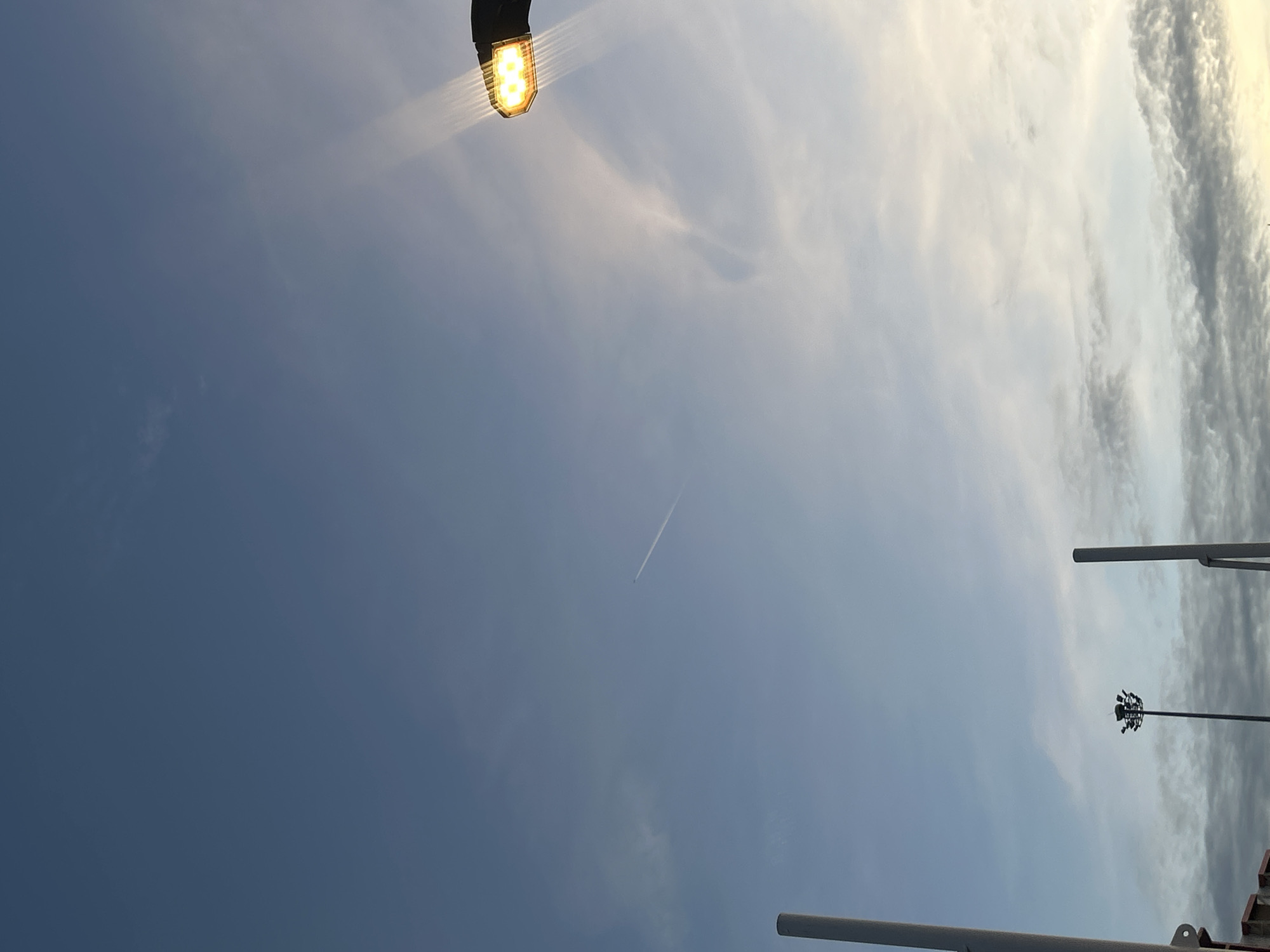 This is a photograph taken outdoors during the day, likely at dusk or dawn, with a clear sky transitioning to a lighter hue near the horizon. The sky occupies the majority of the frame, displaying a gradient of blue with some wispy white clouds. In the upper right quadrant, a streetlamp with a bright yellow light is visible, casting rays of light into the sky. A faint contrail from an airplane stretches across the upper left to middle section of the sky. In the lower portion of the image, the tops of tall poles or light fixtures can be seen. One such fixture is in the center of the frame, a tall pole with multiple lights at its apex. Another tall, slender pole is visible on the far left side of the image. The bottom edge of the image shows a hint of a building or structure, with a red and white patterned element visible. There are no people or animals depicted in the image. The scene suggests an urban or semi-urban environment, possibly a park or a sports field, given the presence of the light fixtures, but the exact location within Tha Khlong Town Municipality, Thailand, cannot be determined from the visual information alone.