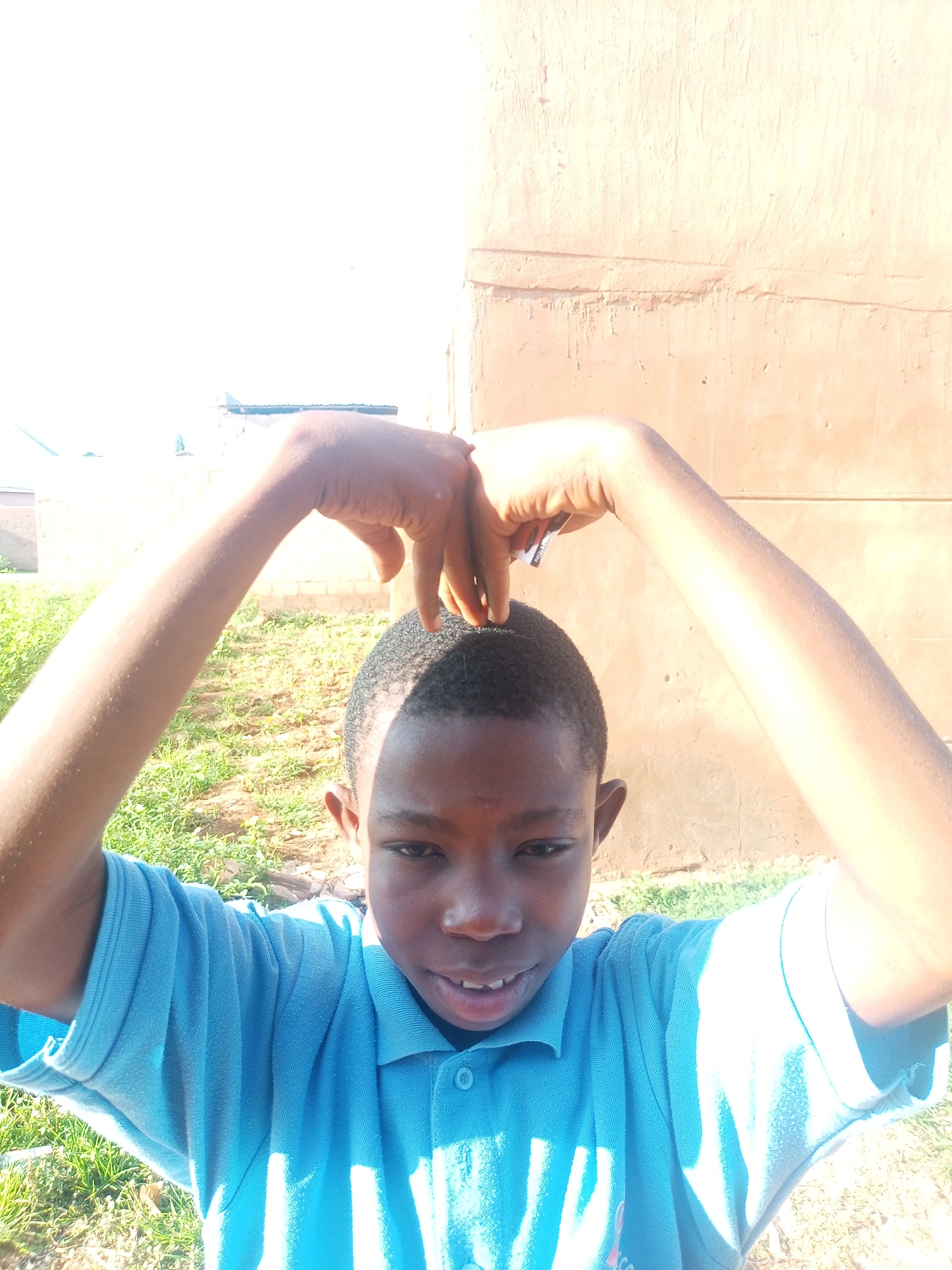 A young boy with dark skin and short, dark hair is posing for a photo outdoors. He is wearing a blue polo shirt. His arms are raised above his head, and his hands are clasped together, forming a heart shape over his head. The boy is smiling. In the background, there is a light-colored, textured wall and green grass. The scene appears to be in a rural or semi-rural area of Tempure, Nigeria.