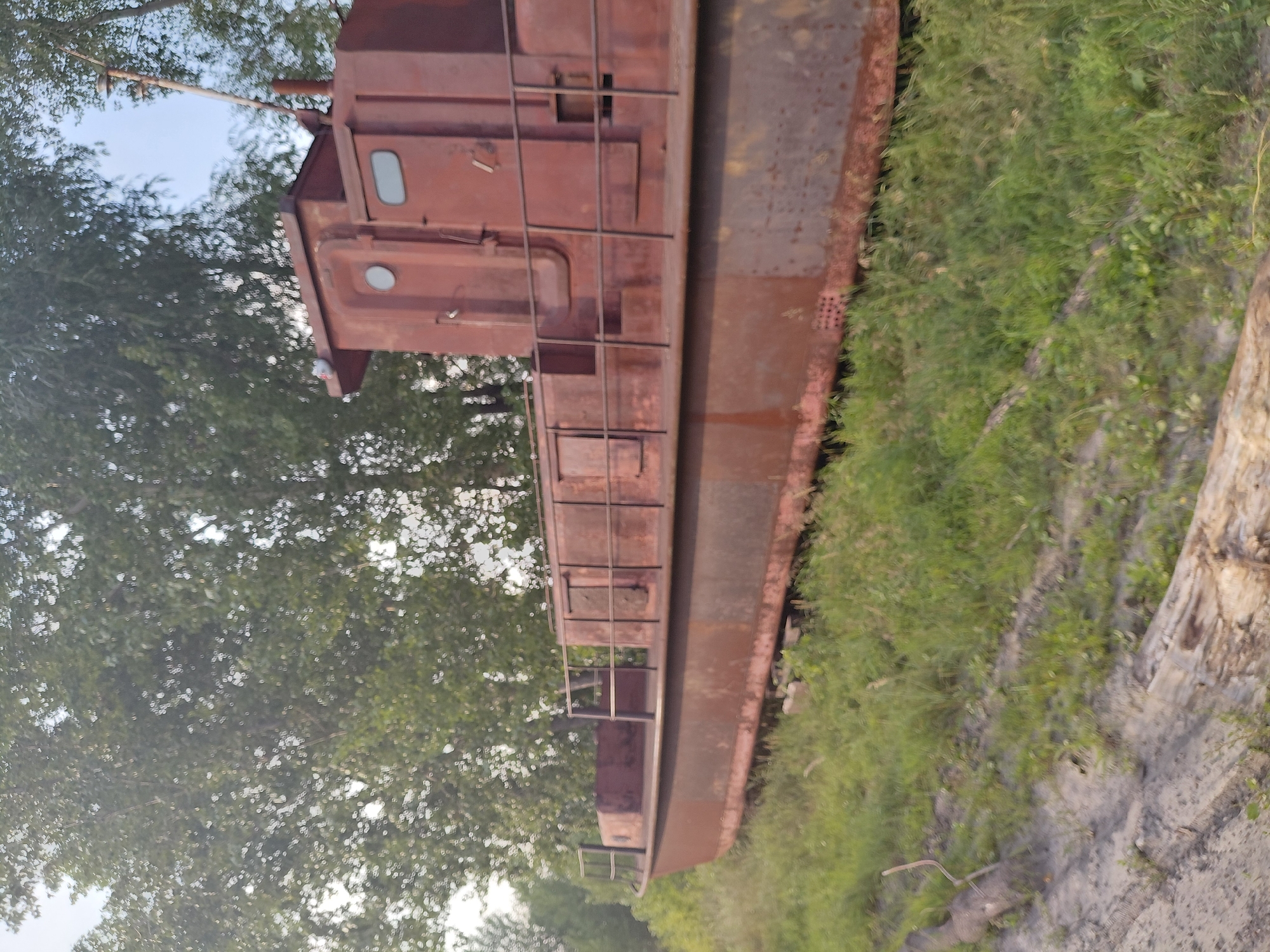 The image shows an old, rusty, red barge beached on a grassy bank. The barge is long and narrow, with a superstructure on the rear half. The superstructure has windows and doors, and a railing runs along the deck. Trees are visible in the background, and the sky is overcast. The scene appears to be in a rural or industrial area. There are no people or animals visible in the image. The barge is likely abandoned. The location is not specified in the image.