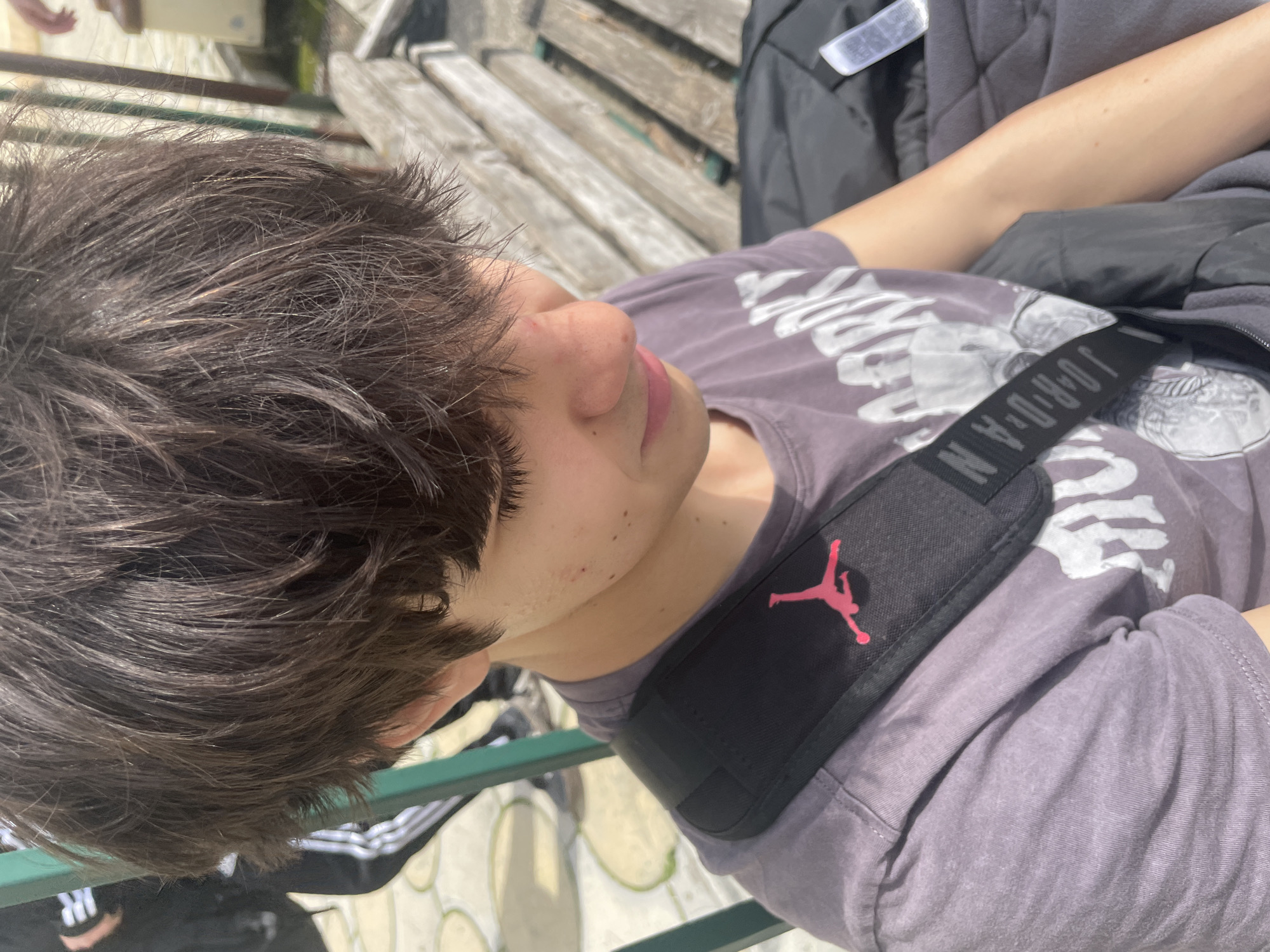 A young person with brown hair lies on a wooden bench. The person wears a grey t-shirt with a white graphic design, and a black Jordan brand messenger bag strap is visible across their shoulder. The setting appears to be outdoors, with patterned paving stones visible in the background. While the city of Varna, Bulgaria, is provided, there are no specific landmarks or details visible to definitively confirm the location.