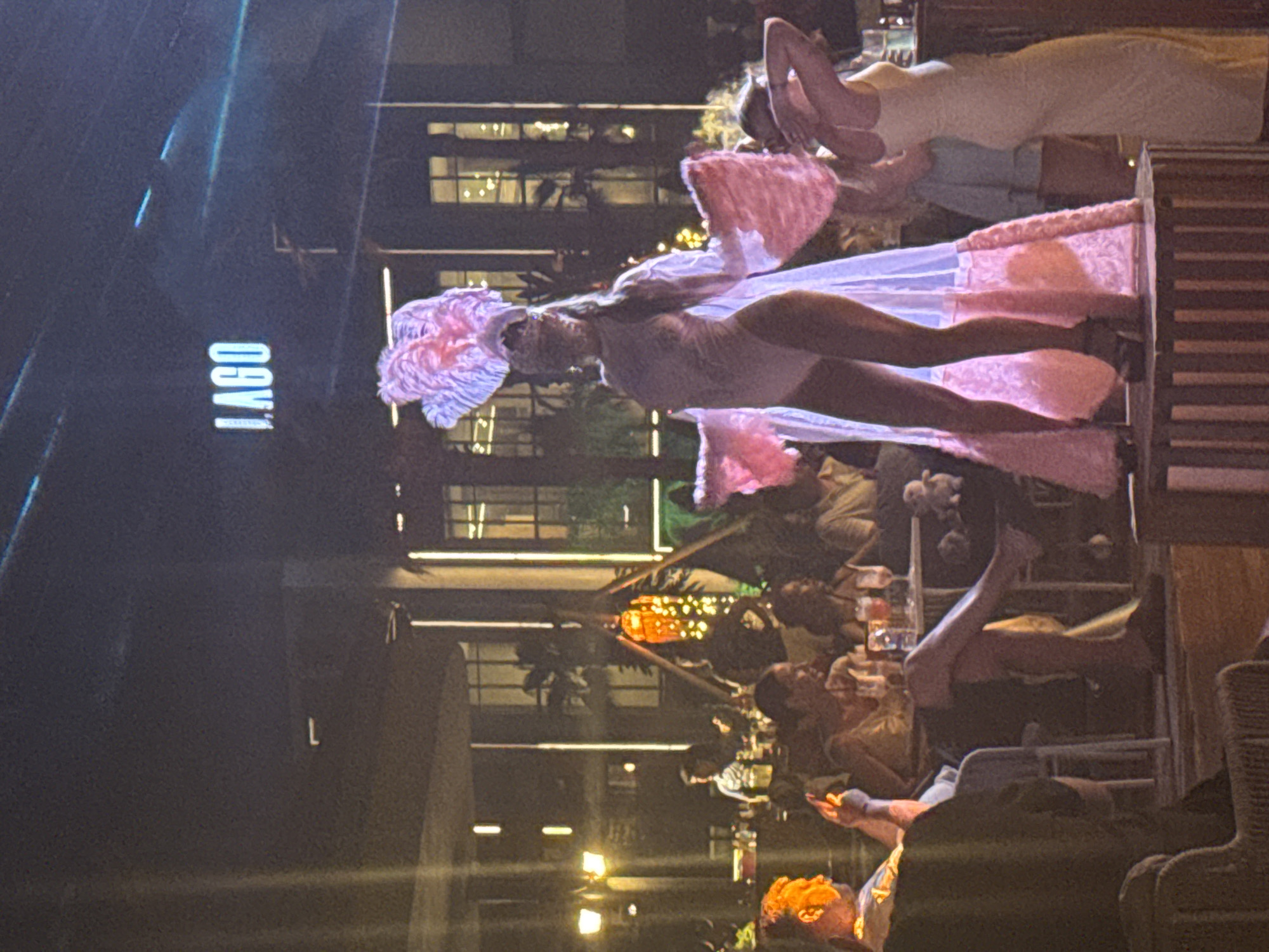 The image depicts an outdoor nighttime scene at a venue with "IAGO" signage prominently displayed. In the foreground, a performer in a light-colored, form-fitting costume and a large, feathered headdress stands on a raised platform. They are also wearing a flowing, sheer cape with pink trim. To the right of the performer, another woman in a pale yellow dress is visible. The middle ground shows a seated audience at tables, with drinks and various objects on them. The background features a building with multiple windows and architectural details, along with palm trees and illuminated decorations, suggesting a lively establishment. The overall atmosphere appears festive and possibly related to entertainment. Given the "IAGO" signage, this venue is likely located in Manavgat, Turkey, known for its tourist attractions and entertainment spots.