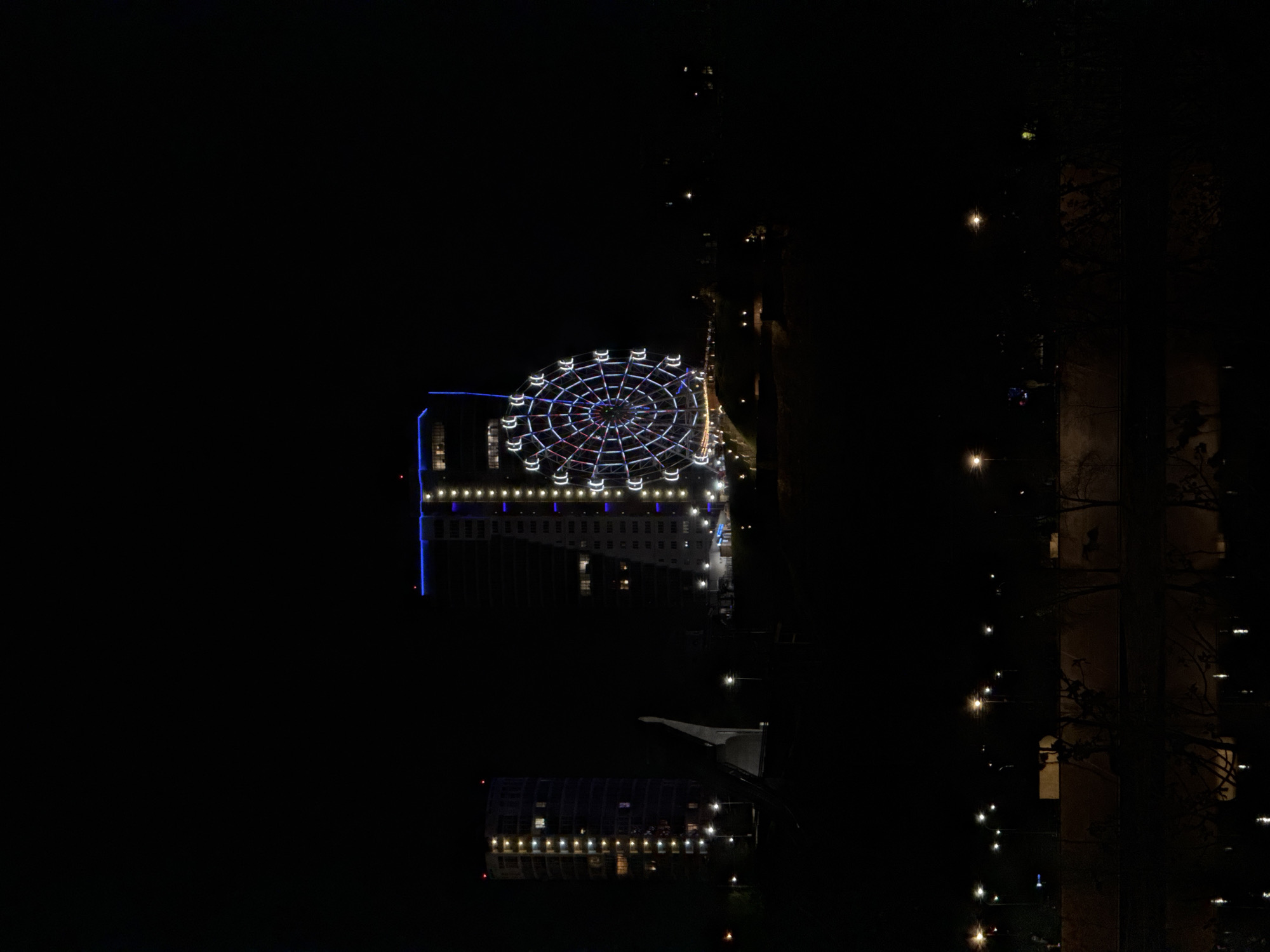 The image shows a nighttime view of a Ferris wheel atop a building in Nizhny Novgorod, Russia. The Ferris wheel is brightly lit with white and blue lights. The building beneath the Ferris wheel is also illuminated. A roadway with streetlights runs alongside the building. A building with streetlights is visible at the bottom of the image. The scene is dark, with limited ambient light. The shot appears to be taken from an elevated position, looking down at the Ferris wheel and building. A dark, out-of-focus building is on the right side of the image.