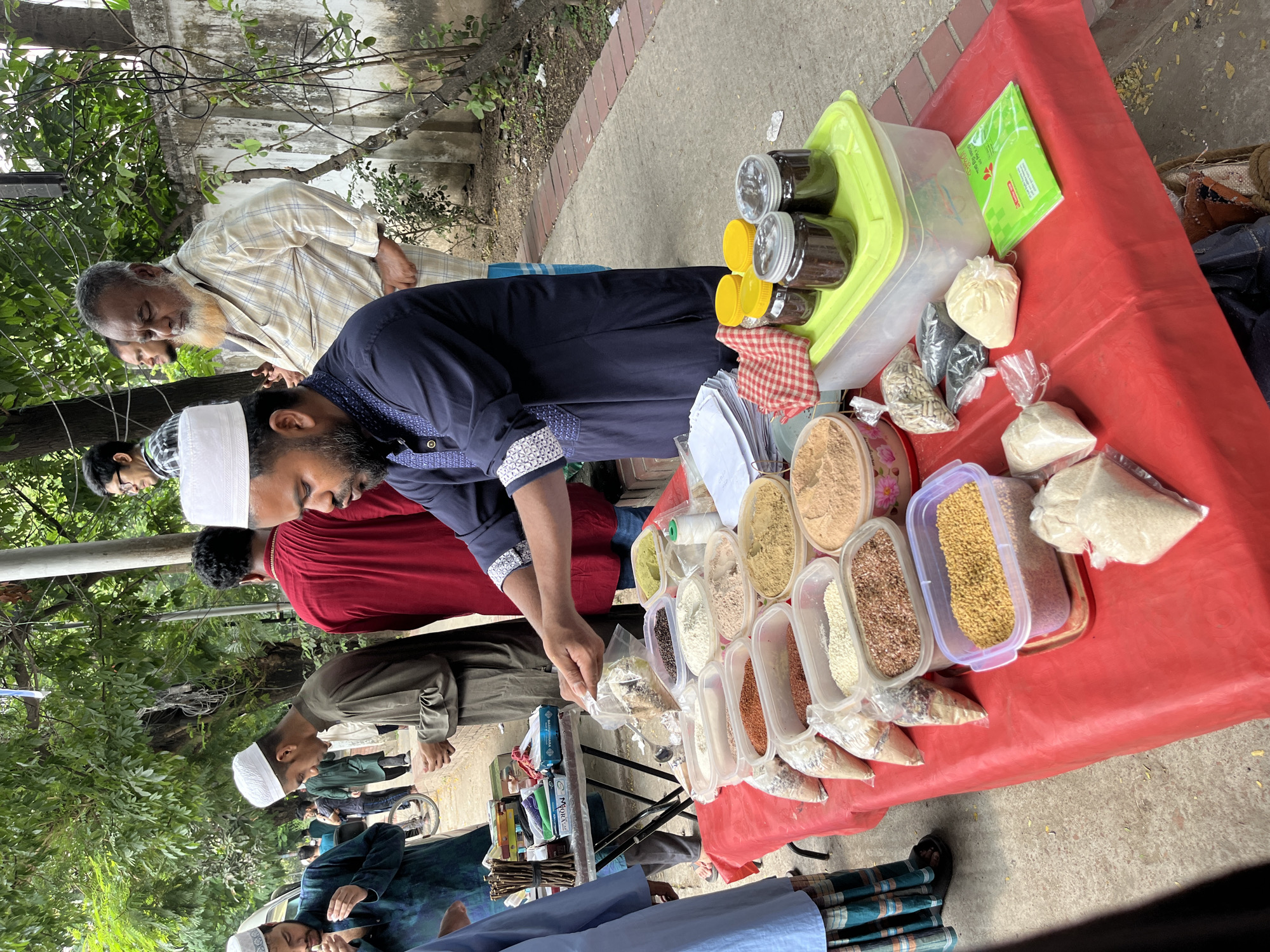 The image depicts an outdoor marketplace scene in Dhaka, Bangladesh. A table covered with a red cloth displays an assortment of spices, grains, and powders in clear plastic containers and bags. A man wearing a white skullcap and a dark blue shirt with patterned cuffs is reaching into one of the containers, presumably to serve a customer. Several other men, also wearing traditional Muslim attire including skullcaps, are standing around the table, likely as customers. Jars and a green box are arranged on the table as well. The location appears to be on a sidewalk adjacent to a street, with trees and a building visible in the background. The sidewalk is made of light gray concrete with a red brick border.