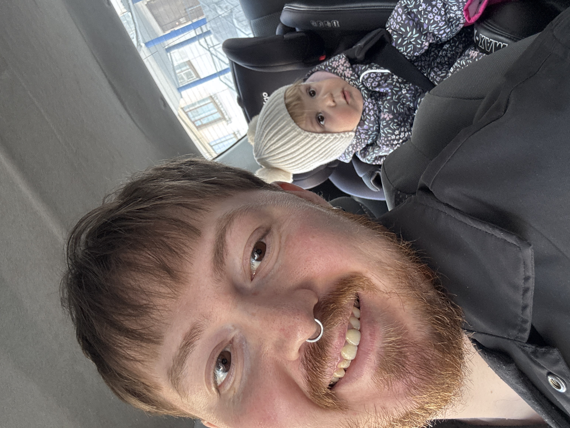 A selfie photograph depicts an adult individual with brown hair and a red beard, smiling into the camera while inside a vehicle. The individual is wearing a dark-colored jacket and has a ring in their nose. Behind the individual, a baby wearing a white knitted hat with a pom-pom and a floral-patterned jacket is secured in a black car seat. Through the car window, a glimpse of buildings is visible, potentially suggesting an urban environment. Based on the building architecture and window designs, the location is potentially Chaikovsky, Russia. The action is a selfie being taken inside a vehicle with a child present. The environment appears to be the interior of a car during daylight hours.