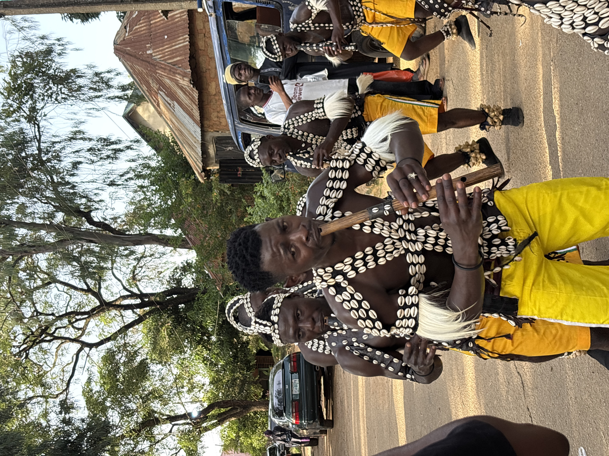 The image shows a group of men in traditional attire participating in what appears to be a cultural event or procession in Jos, Nigeria. The men are wearing black and white beaded vests and headwear adorned with cowrie shells and white feathers. Several of them are playing on wooden flutes. One man in the foreground is holding a flute to his lips, his hands positioned on the instrument. Another man behind him is also playing a flute. A few other individuals are visible behind them, including one in a white t-shirt with text that appears to read "Teamewer."
They are dressed in bright yellow shorts or pants, contrasting with their upper garments. Some of the men have anklets made of shells that jingle with movement. The scene takes place outdoors, on a paved road. In the background, there is a building with a corrugated iron roof, and lush green trees line the side of the road. A blue vehicle is parked nearby, and further down the road, other cars are visible. The lighting suggests it is daytime, likely late afternoon or early evening, given the warm, golden hues cast by the sun. The overall atmosphere seems festive and celebratory, with a focus on cultural expression and music.