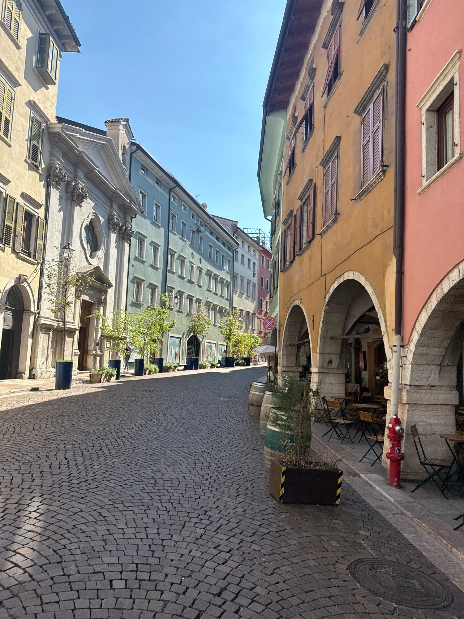 The image captures a view down a cobblestone street in Trento, Italy, on a sunny day. The street is lined with historic buildings displaying a variety of pastel colors, including pale yellow, mint green, and salmon pink.
On the left side of the street, a light yellow building with shutters is visible, next to a church-like structure with a grand facade and an arched entrance. Several potted trees with green foliage are placed along the sidewalk in front of the buildings. Further down the street, more colorful buildings continue, some with arched windows.
On the right side of the street, a building with a pale orange facade features a series of stone arches. Beneath these arches, outdoor seating with tables and chairs is set up, suggesting a cafe or restaurant. A bright red fire hydrant stands near the arches. Several wooden barrels and planters are also present along the street. The sky is a clear blue, and shadows are cast across the cobblestone pavement. A circular manhole cover is visible on the ground towards the right side of the image. A single person is seen walking in the distance on the left side of the street.
