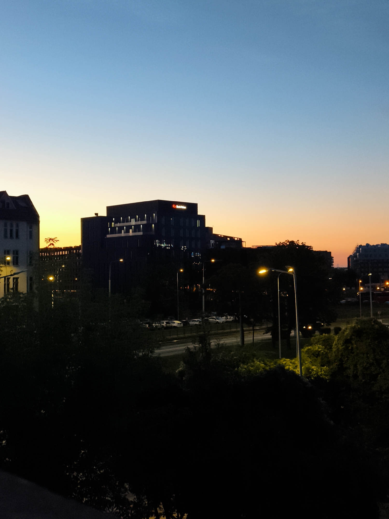 The image shows a cityscape at dusk, likely in the city of Gdańsk, Poland. The sky transitions from a clear, pale blue at the top to a warm orange and yellow gradient near the horizon, indicating the setting sun. Silhouetted against this colorful sky are several buildings. On the left, a lighter colored, older building with a pitched roof is visible. To its right, a darker, modern building with multiple floors and illuminated windows stands prominently. Further to the right, another dark building with angular architecture is present. A street with a few cars is visible in the middle ground, with streetlights casting a warm glow. Trees and foliage are in the foreground, mostly in shadow, creating a dark silhouette that frames the scene. The overall impression is one of an urban environment at twilight.