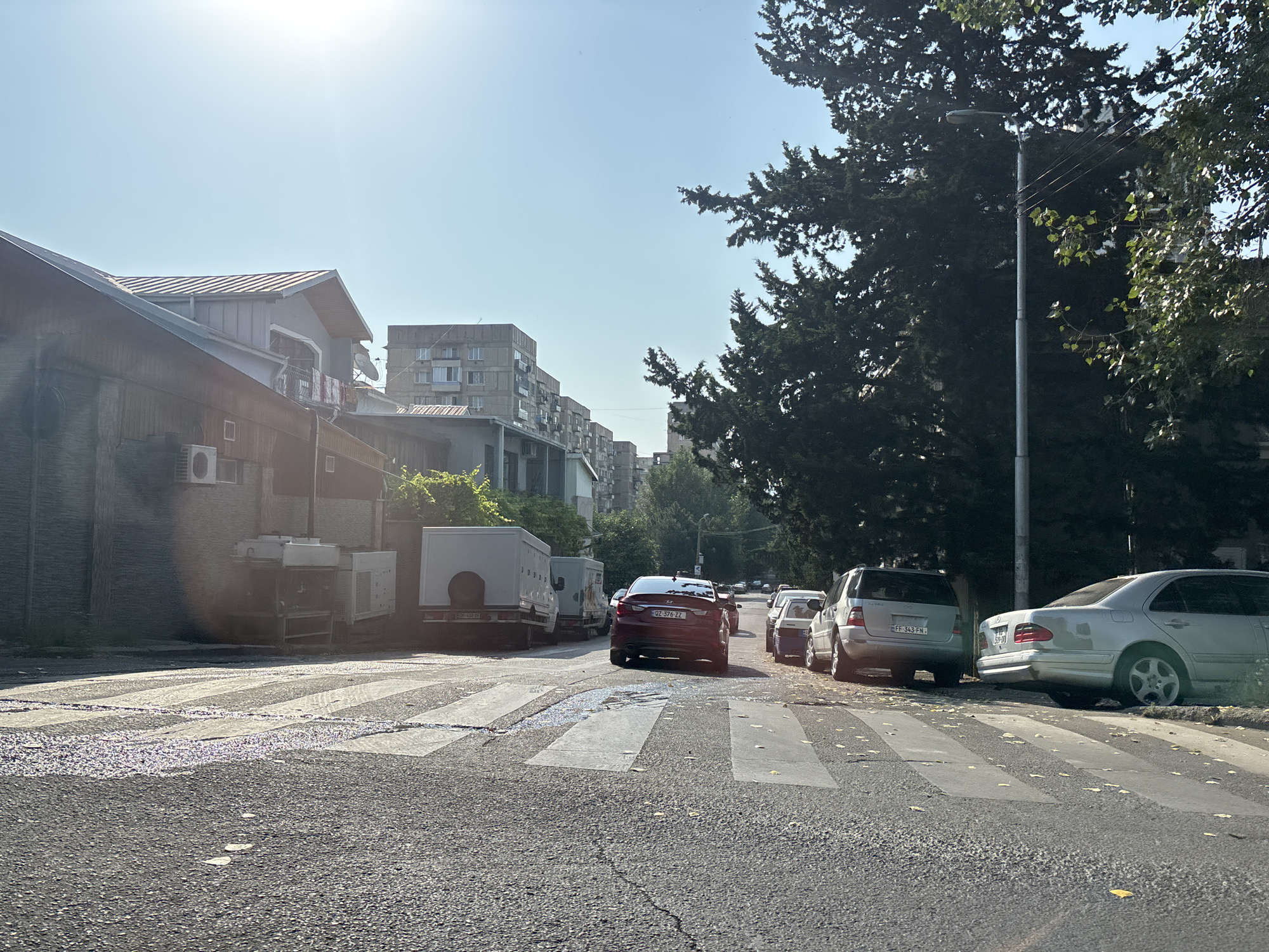 The image depicts a street scene in Tbilisi, Georgia. In the foreground, a cracked asphalt road features a pedestrian crossing with white stripes. A dark red sedan with license plate "UZ-376-ZZ" is positioned on the crossing, facing away from the camera. To the right of the sedan, parked cars are visible: a silver SUV with license plate "FF-343-FN" and a silver sedan. In the background, a large coniferous tree dominates the right side of the frame. On the left, buildings include a lower structure with an air conditioning unit and a taller, multi-story apartment building. The sky is clear and bright, indicating daylight. The scene appears to be an ordinary urban environment.