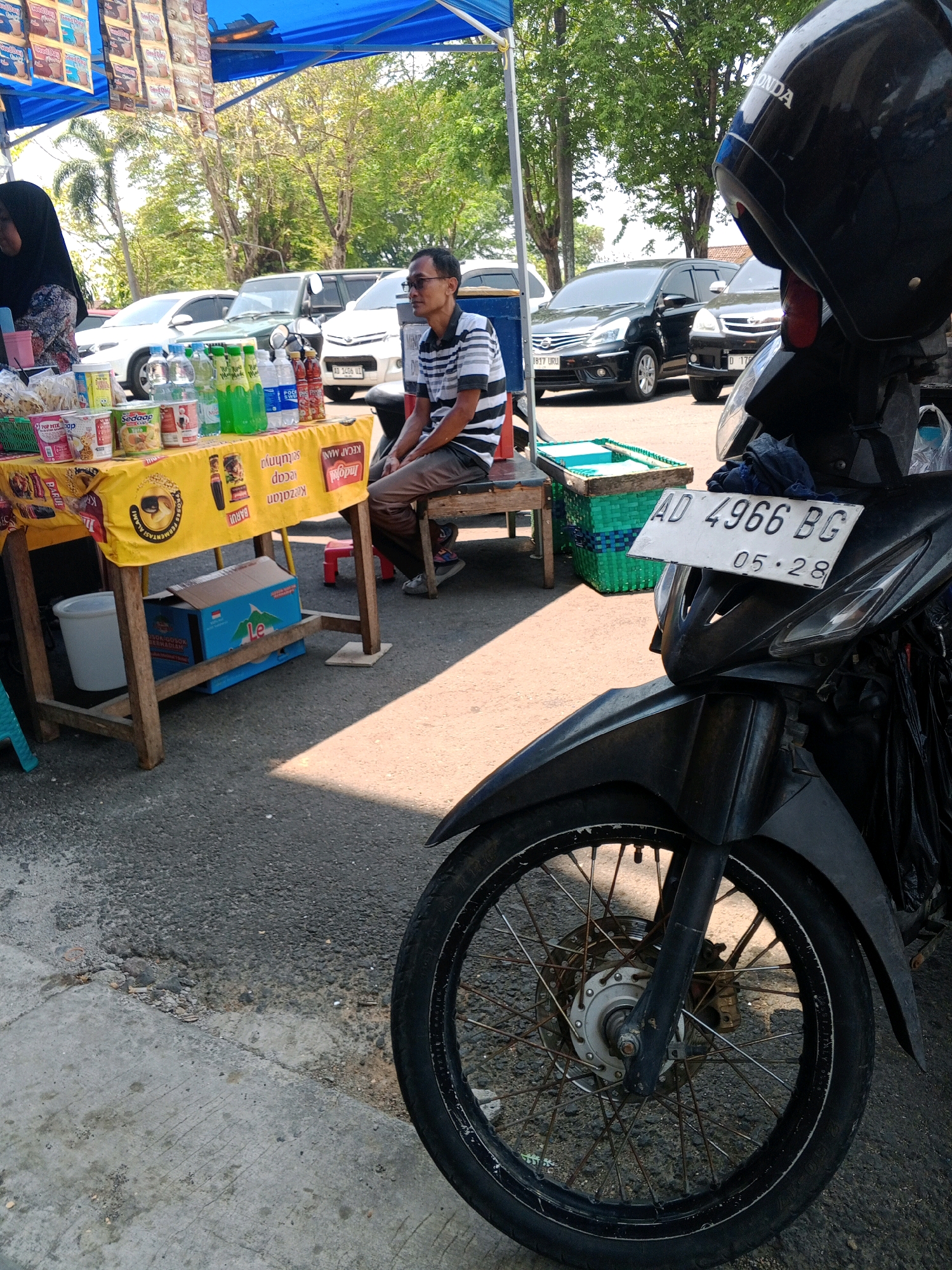 A man is sitting on a bench next to a table filled with goods. The table is covered with a yellow cloth and has various items like drinks, snacks, and sauces. Behind the table, several cars are parked. In the foreground, a motorcycle is parked, partially obscuring the view. The scene appears to be an outdoor market in Wuryorejo, Indonesia, given the presence of local products and Indonesian license plates on the vehicles. The man is wearing a striped shirt and glasses, and he is looking towards the right of the frame. The overall atmosphere suggests a typical day at a roadside market.