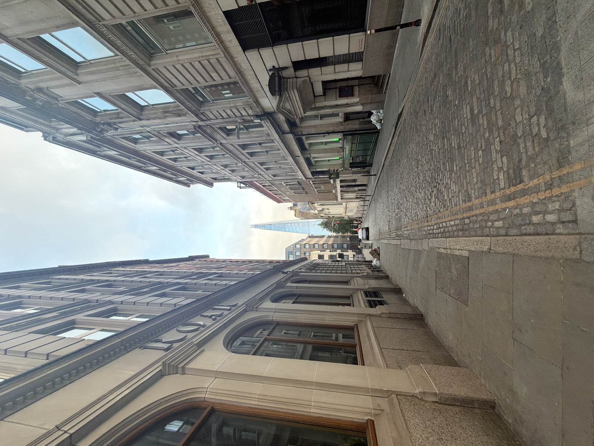 This photograph captures a street scene in London, United Kingdom. Tall buildings line either side of the street, their architecture displaying historical features such as arched windows, stone facades, and multiple floors of windows. The street is paved with cobblestones, marked by yellow lines. A vehicle is parked further down the street. The Shard is visible in the distance, piercing the sky between the buildings. The sky is overcast.