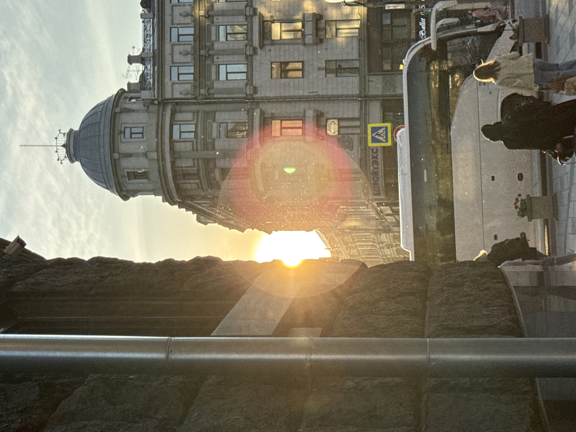 The image, taken in Moscow, Russia, shows a sunny urban scene. An elaborate stone building with a distinctive dome occupies the upper left portion of the frame. In the upper right, a bus is partially visible, with several people standing near it, including one in a light-colored jacket and others in darker attire. The sun is positioned between the building and the foreground, creating a lens flare effect. The foreground consists of rough stone elements and a metal pipe running horizontally across the bottom of the image.