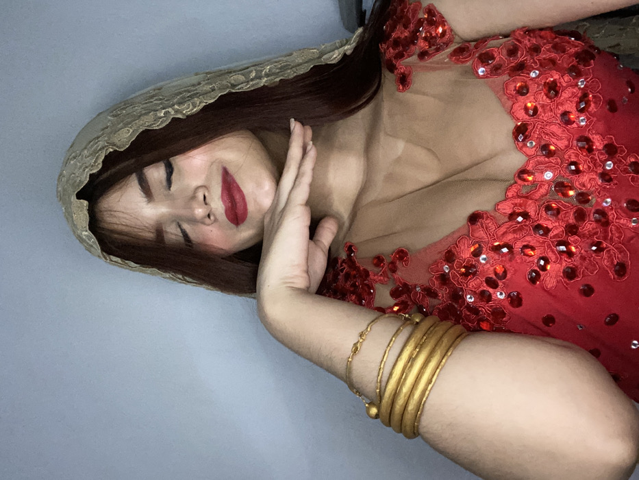 A woman in a red dress with ornate beading poses with a lace-edged, gold-toned headscarf. She wears gold bangles on her arm, which is raised towards her face. Her eyes are closed, and she wears dark lipstick and rosy blush. The background is a pale gray.