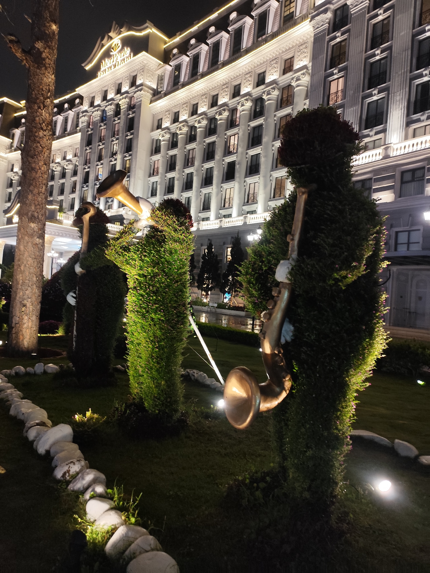 The image presents a captivating nighttime view of a hotel garden featuring whimsical topiary art. In the foreground, three distinct topiary figures, meticulously sculpted from dense green foliage, resemble musicians playing brass instruments. The figure on the right holds a golden saxophone, while the central figure appears to play a golden trombone or trumpet, with white glove-like hands visible. A third figure is partially obscured by a large tree trunk on the left.
The topiaries and parts of the lush green lawn are illuminated by discrete ground spotlights, creating a vibrant glow. A decorative border of white, rounded stones lines the edge of the garden bed in the lower left.
Dominating the background is a large, stately multi-story building, brightly lit against the dark sky. Its architecture is grand and ornate, characterized by numerous windows, classical columns, elaborate cornices, and decorative balconies. The building's roofline and architectural details are accentuated by strings of warm white lights, highlighting its majestic presence. Prominently displayed on the building's facade, beneath an emblem, is the text "MerPerle Dalat Hotel." The setting is an elegant outdoor space at night, suggesting a luxurious and inviting atmosphere.
