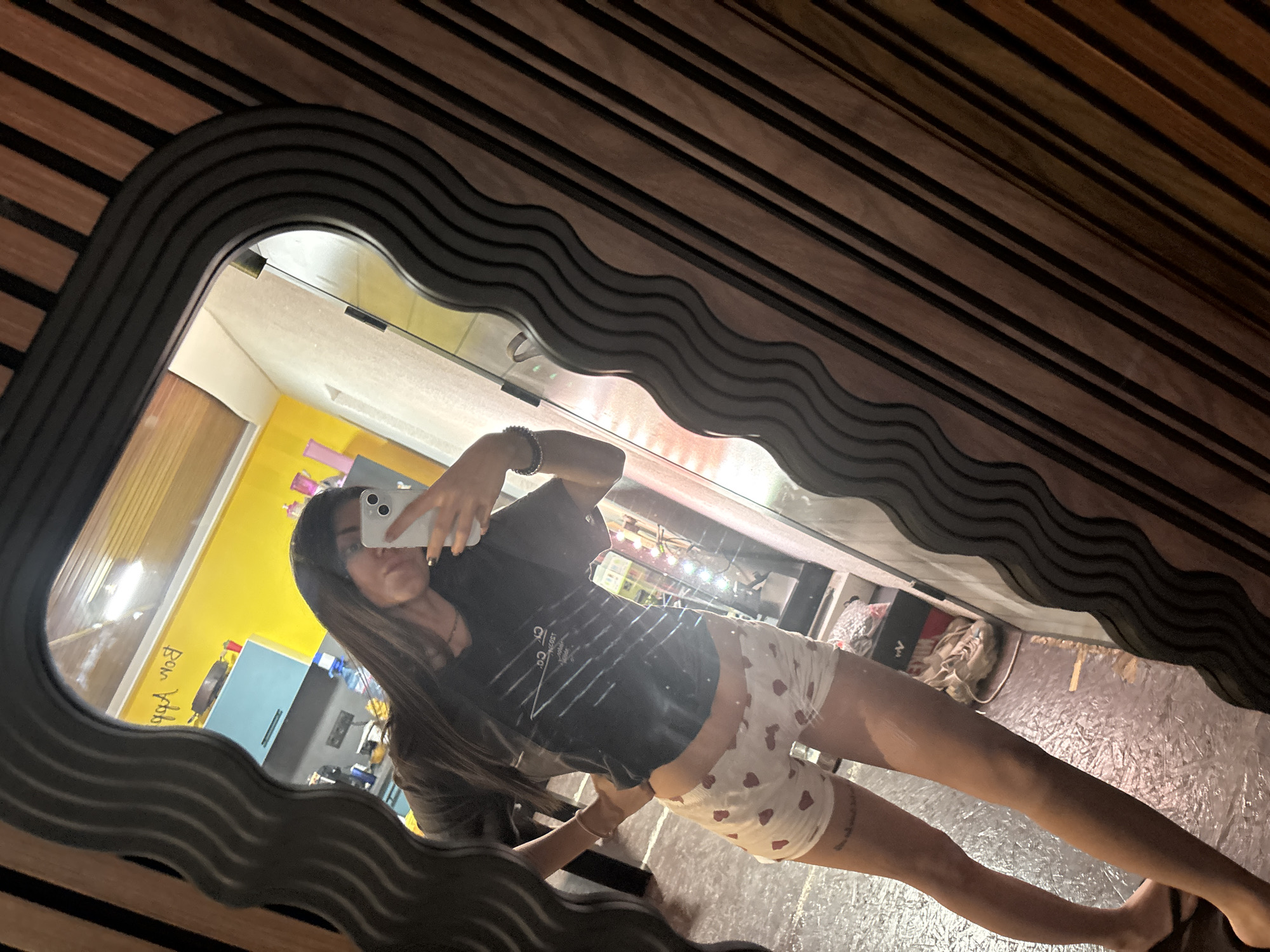 The image shows a selfie taken in a mirror with a wavy frame. The individual in the reflection is a young woman holding a smartphone, taking the photo. She's wearing a dark-colored t-shirt and patterned shorts. The reflection reveals a room with a yellow wall, a blue cabinet, and what appears to be kitchen appliances. The floor is tiled. Outside the room, there are shoes and a bag. The location cannot be determined as Belopoptsi, Bulgaria from the provided information.