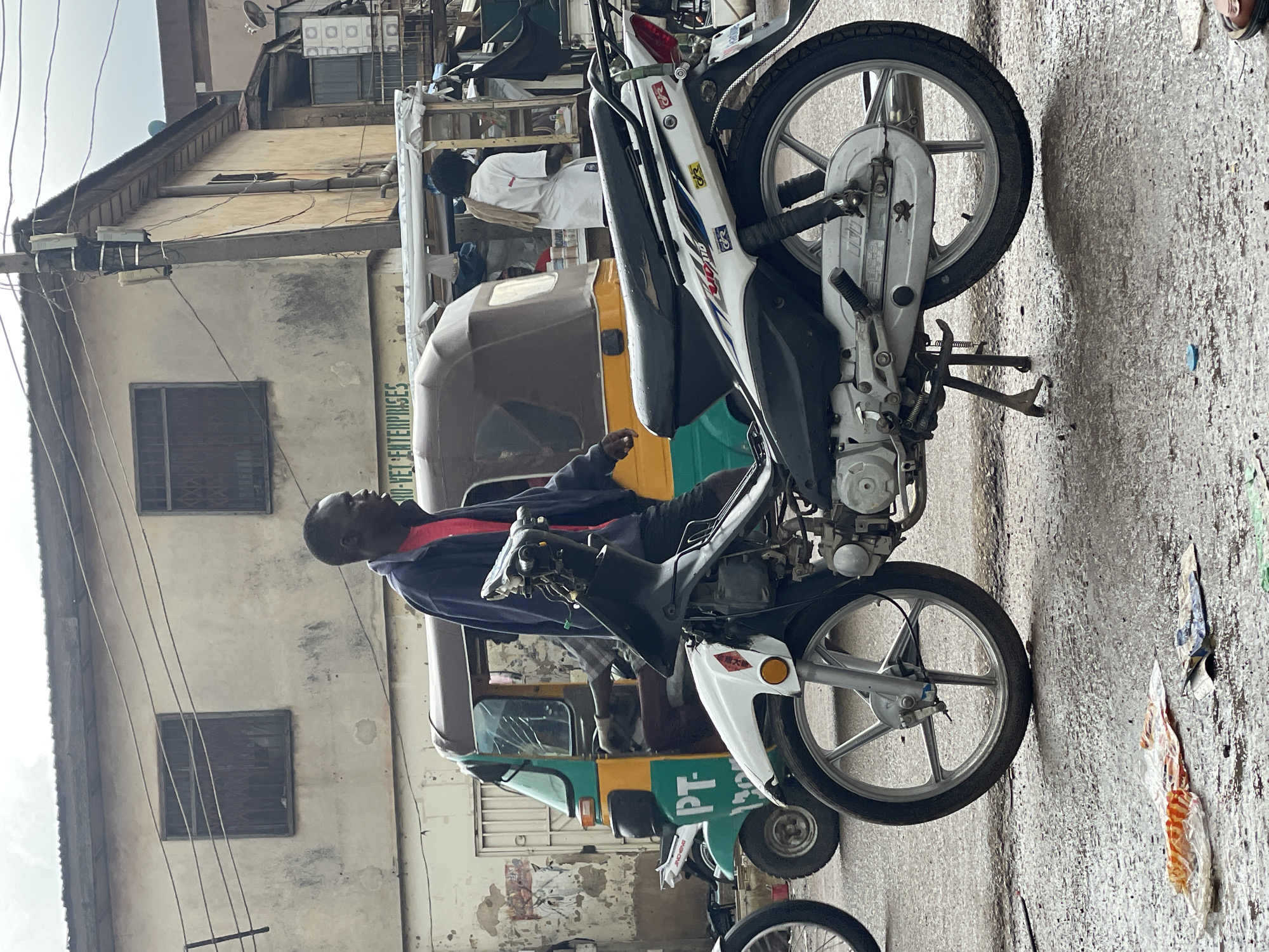 A man sits on a white and black motorcycle parked on a street in Jos, Nigeria. He is wearing a blue jacket over a red shirt and appears to be looking upwards. The motorcycle is a common model with a silver frame and black tires. Behind the motorcycle, a yellow and green auto-rickshaw is partially visible, suggesting a busy urban environment. The buildings in the background are weathered and have visible windows and electrical wires. The ground is paved with rough concrete, and there are some pieces of debris scattered around. The lighting suggests it is daytime, with bright sunlight casting shadows. The overall scene depicts everyday life in a Nigerian city, with transportation and local businesses being prominent. The image includes the text "PT" and "VEP ENTERPRISES" on the side of the auto-rickshaw.