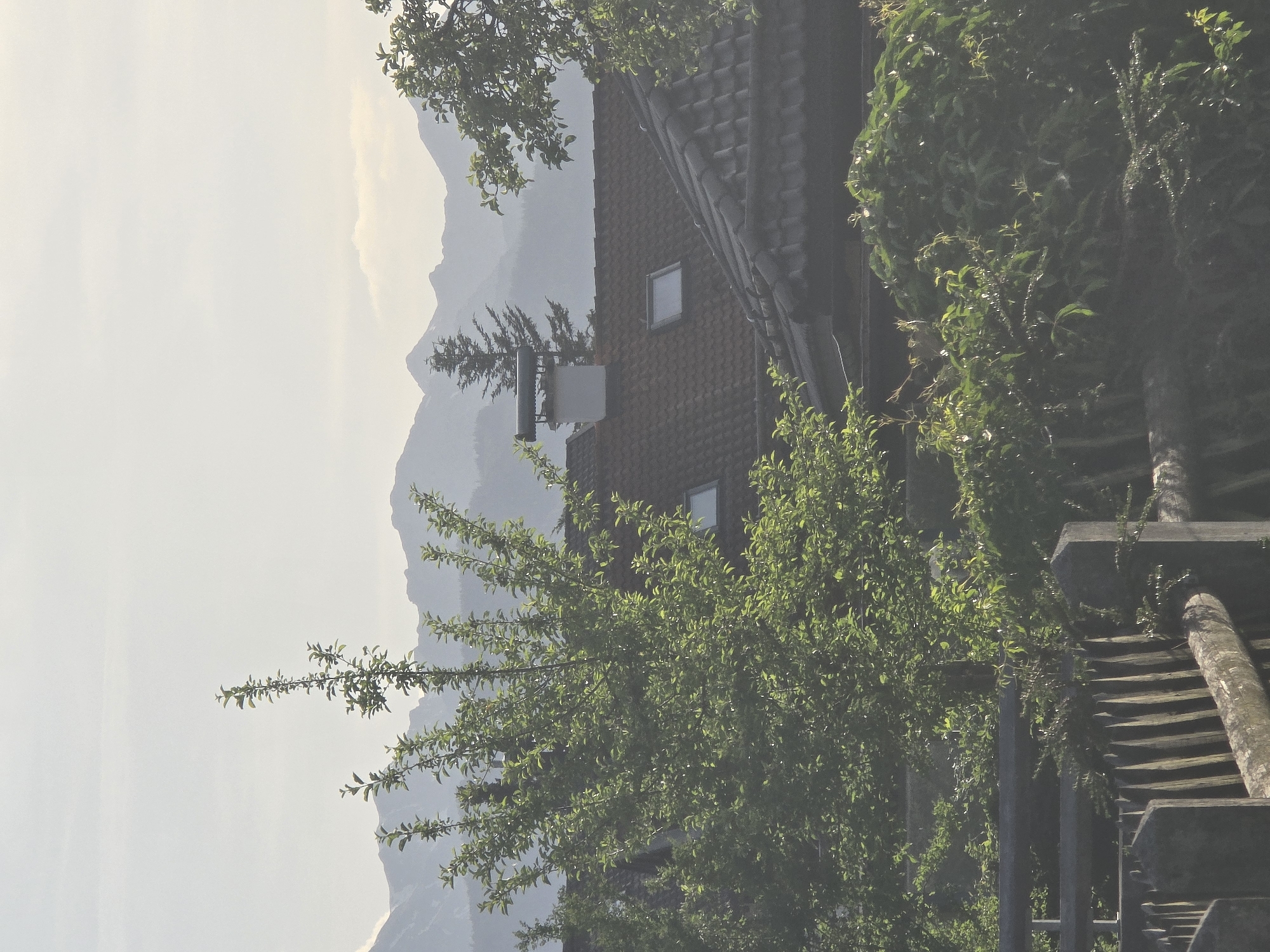The image captures a mountainous landscape with a house nestled among trees. The house has a reddish-brown roof with small, square windows. A chimney or antenna is visible on the roof. Lush green foliage surrounds the house and partially obscures the view. In the background, hazy mountains rise, blending into the sky. In the foreground, a wooden structure with planks and supports is visible. Based on the landscape and architectural style, the image likely depicts a scene in or near Garmisch-Partenkirchen, Germany, a region known for its mountains and traditional Bavarian architecture.