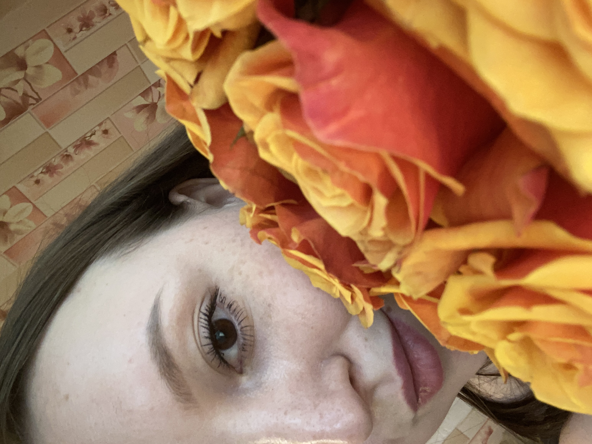 The image is a close-up of a young woman holding a bouquet of orange and yellow roses. She is partially visible, with her left eye, nose, and lips in focus. The roses obscure a large portion of her face and the background. The background appears to be a wall with a floral wallpaper pattern. The lighting suggests an indoor setting, possibly a home. There is no indication of the city of Chaikovsky, Russia, or any specific location. The woman is looking towards the camera with a neutral expression. Her skin has freckles. The roses are vibrant and in full bloom.