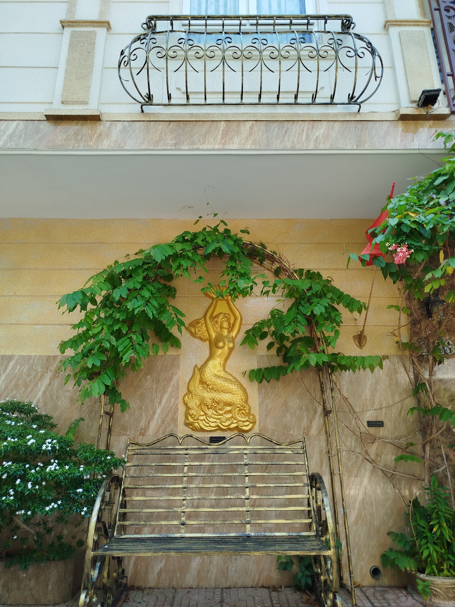 A medium shot shows a golden mermaid sculpture mounted on a tan, marble-patterned exterior wall above a wrought iron and wood bench in Ho Chi Minh City, Vietnam. Lush green vines climb the arched metal frame around the mermaid, nearly obscuring it. To the left, a flowering plant with small white flowers adds a touch of nature. To the right, another vine with red flowers adds color. Above the wall, an iron balcony with a decorative design adds architectural interest. The environment suggests a blend of art, nature, and residential architecture.