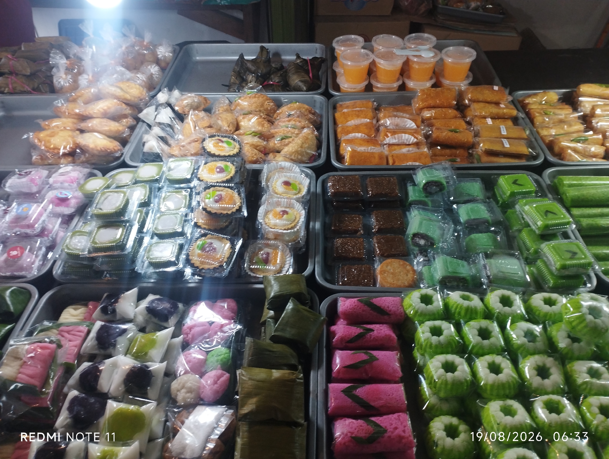 A wide shot captures an outdoor market stall in Karawang, Indonesia, displaying a variety of Indonesian snacks and desserts. The stall is organized into several rectangular trays, each filled with individually packaged food items.
In the background, a tray holds several banana leaf-wrapped parcels, possibly containing rice or savory dishes. To the right of these, another tray features small plastic cups filled with orange-colored liquid, likely a beverage or dessert.
The middle section of the stall is a panorama of sweet treats. One tray is filled with numerous golden-brown, fried pastries, many of which appear to be "risoles" or similar fried snacks. Next to these, a tray contains rows of orange cake slices, some wrapped in paper. Further to the right, more fried pastries are visible.
In the foreground, several trays showcase a colorful assortment of desserts. One tray holds small tartlets with fruit toppings. Another tray displays small green and white square-shaped cakes, some with decorative green markings. Adjacent to these are rows of bright pink, rolled cakes, also with green leaf decorations. A tray in the lower left corner is packed with various small, colorful steamed cakes and sweet treats in white and purple wrappers, alongside some pale pink, flower-shaped cakes.
The overall lighting is bright, with a strong light source from the upper left, creating some glare on the plastic packaging. The market appears busy, though no people are clearly visible in the frame, suggesting the photograph was taken from a customer's perspective. The stall itself is likely a temporary setup at a local market or roadside, a common sight in Indonesian cities like Karawang. The timestamp "19/08/2026. 06:33" and "REDMI NOTE 11" are overlaid in the bottom right and left corners, respectively, indicating the photograph was taken on a specific date and time using a mobile phone.