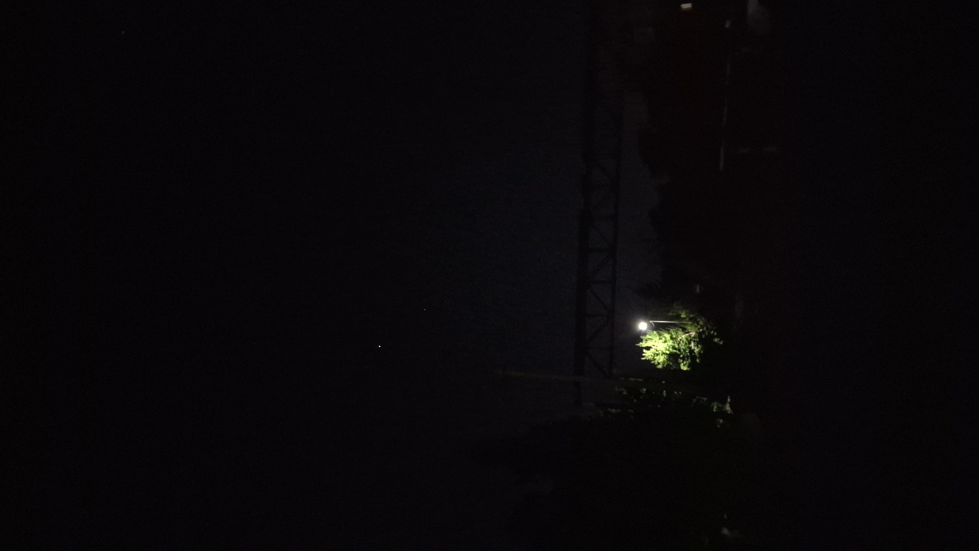 A night-time, low-light photograph captures a distant industrial bridge spanning across the frame, its metallic structure silhouetted against the dark sky. Below the bridge, a single streetlamp illuminates a patch of vibrant green foliage, creating a stark contrast with the surrounding darkness. Two faint celestial bodies are visible in the upper portion of the image, appearing as small, bright dots against the inky blackness of the night sky. The overall scene is one of urban infrastructure blending with the natural elements under the cloak of night. The image is taken in Aksu City, China.