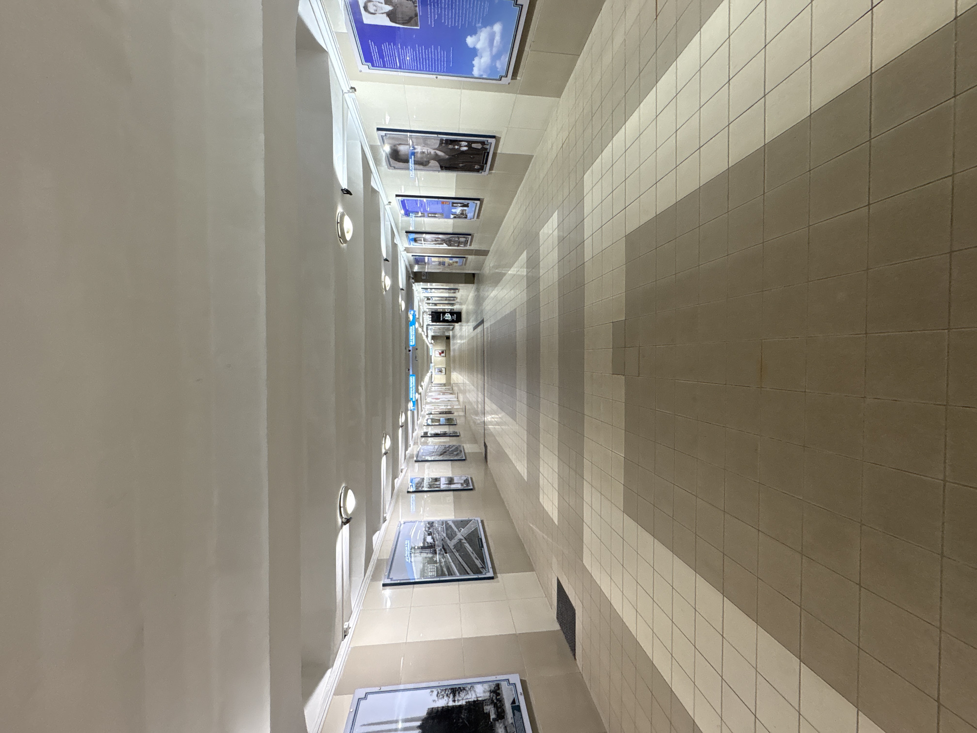 The image shows a long, well-lit underground pedestrian tunnel. The tunnel has a tiled floor with a pattern of light and dark beige tiles. The walls are also tiled, with a beige and cream color scheme. Along the walls, there are framed displays featuring historical photographs and portraits. Several overhead lights illuminate the tunnel. At the far end of the tunnel, there are signs and a vending machine. The scene appears to be clean and well-maintained, suggesting it is a public space in regular use. This tunnel is located in the city of Vitsebsk, Belarus, and is likely a pedestrian underpass connecting different parts of the city.