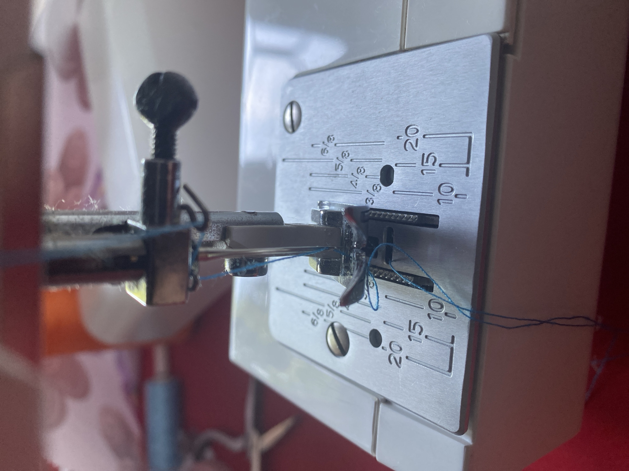 This is a close-up, overhead shot of a sewing machine needle and presser foot with blue thread. The needle is lowered and threaded through the eye of the presser foot, with excess thread trailing down. The presser foot is positioned over the metal stitch plate of the sewing machine, which has inch and centimeter markings. The background is blurred and shows glimpses of other sewing supplies, including spools of thread and scissors. The scene appears to be indoors, likely in a home or workshop setting. There are no people or animals visible in the image, and no specific location in Orlivshchyna, Ukraine can be identified from the visual information provided.