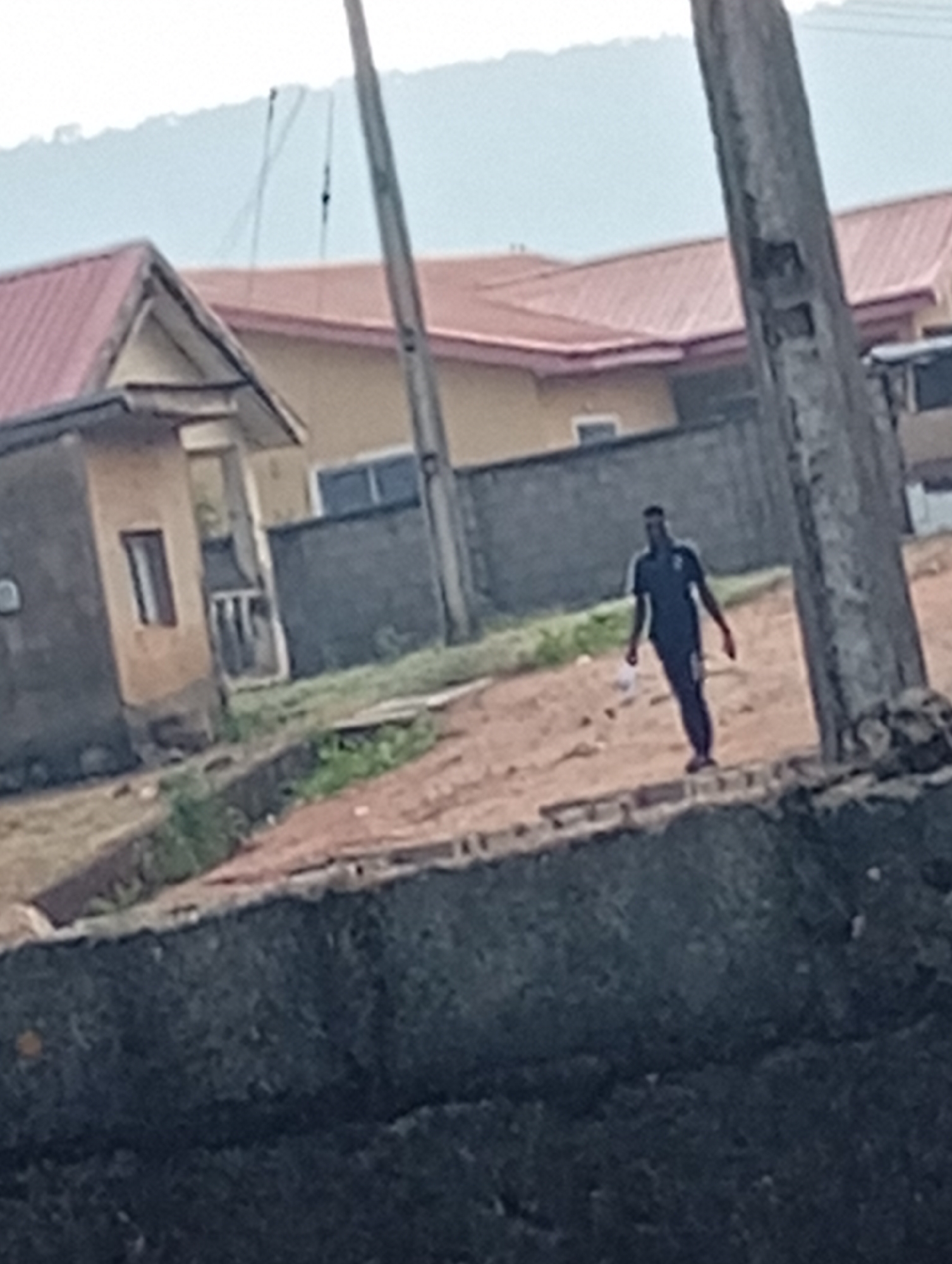 A street scene in Adankolo, Nigeria. A man in a dark tracksuit walks on a dirt road near a concrete wall. In the background, there are houses with red and orange roofs and power lines are visible. The weather is overcast.