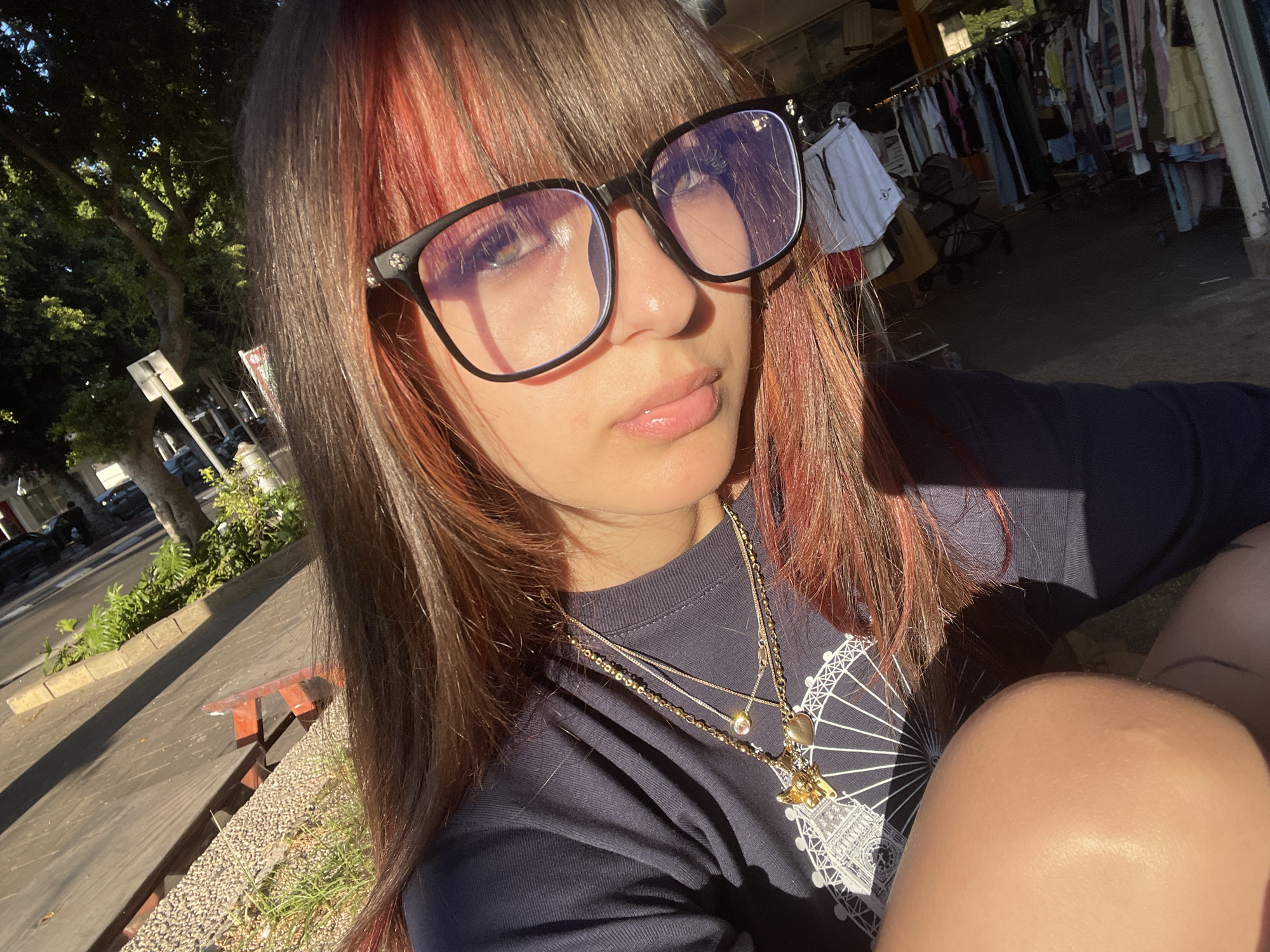 A young person with long, dark brown hair with pink streaks and bangs is shown in a close-up, outdoor selfie. They are wearing black-rimmed glasses and a dark t-shirt with a white graphic of what appears to be the London Eye. Multiple gold necklaces are layered around their neck, with pendants including a heart, a small jewel, and a charm. The person is looking slightly to their left, with their lips slightly parted.
The background suggests a public outdoor setting, possibly a market or street. In the upper right, racks of clothing are visible, hinting at a market stall. To the left, a street with trees and sidewalk is visible, with cars parked in the distance. The lighting suggests it is daytime, with sunlight casting shadows and highlighting the person's face and hair. The overall emotion conveyed is relaxed and casual. The location is identified as Raanana, Israel.
