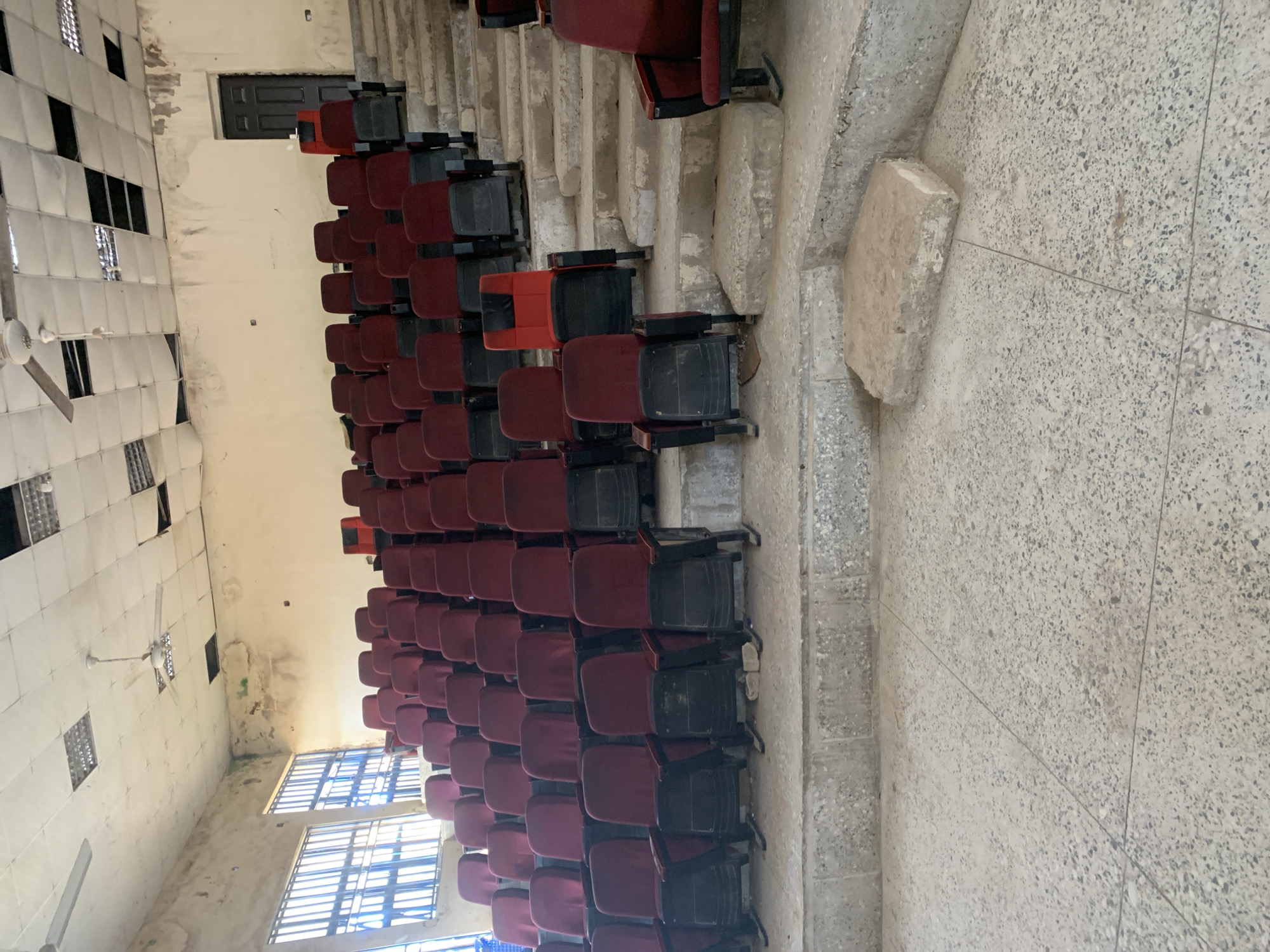 The image shows an interior view of an auditorium or lecture hall with tiered seating. The seating consists of burgundy and black chairs with metal frames. The room appears to be in a state of disrepair, with a damaged ceiling featuring exposed beams and missing tiles, water stains on the walls, and cracked concrete steps. There are large windows with security bars on the left side of the room, and a dark-colored door is visible in the background on the right. The floor is concrete with visible seams. A single red chair stands out among the burgundy ones. The scene is devoid of people. This location is the auditorium of the Federal College of Education, Jalingo, Nigeria.