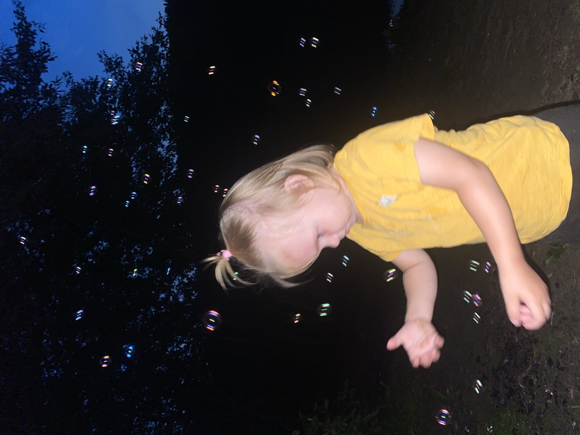 A young Caucasian child with blonde hair, wearing a yellow t-shirt, is in the process of blowing bubbles outdoors in what appears to be a park or natural area at dusk. The child's face is partially visible as they look down and to the right, with their left hand outstretched as if to catch or interact with the bubbles. The background is dark, with silhouettes of trees against a deep blue twilight sky. Numerous colorful, iridescent bubbles are floating in the air around the child, reflecting light. The ground is dark and appears to be covered with dirt or mulch, with some foliage visible on the left side of the frame. The overall scene suggests a recreational activity taking place in a natural setting during the evening hours. The location is identified as Novosibirsk, Russia.