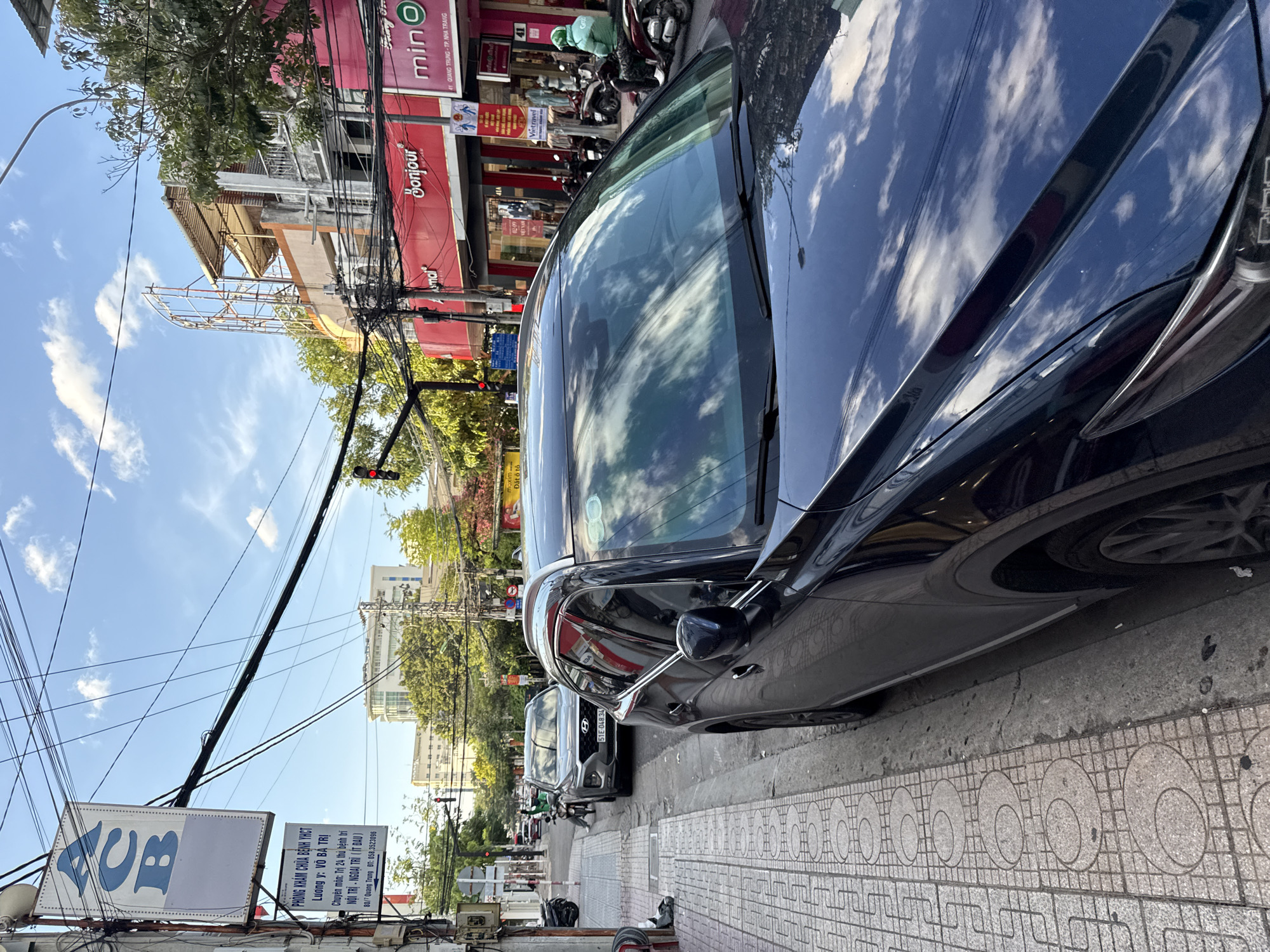 A dark blue SUV is parked on the right side of a street in Nha Trang, Vietnam. The vehicle is in the foreground and takes up a significant portion of the right side of the image. The SUV's windshield reflects the sky and the surrounding buildings.
To the left of the SUV, a sidewalk with a patterned tile design is visible. Beyond the sidewalk, a gray sedan is parked. Further down the street, a motorcycle with a rider wearing a green helmet is approaching.
On the left side of the street, a pole with several signs is mounted. One sign is white with blue lettering that spells out "ACB". Below it, a smaller white sign with black text provides information about a medical clinic: "PHÒNG KHÁM CHỮA BỆNH YHCT", "LƯƠNG Y VÕ BÁ TRÌ", and an address.
Across the street, buildings line the sidewalk. One building has a prominent red facade with the word "Bonjour" in white lettering. Another sign reads "min O". Various shops and storefronts are visible, and several motorcycles are parked along the street. Overhead, a complex network of electrical wires is strung across the sky. The sky is blue with scattered white clouds.