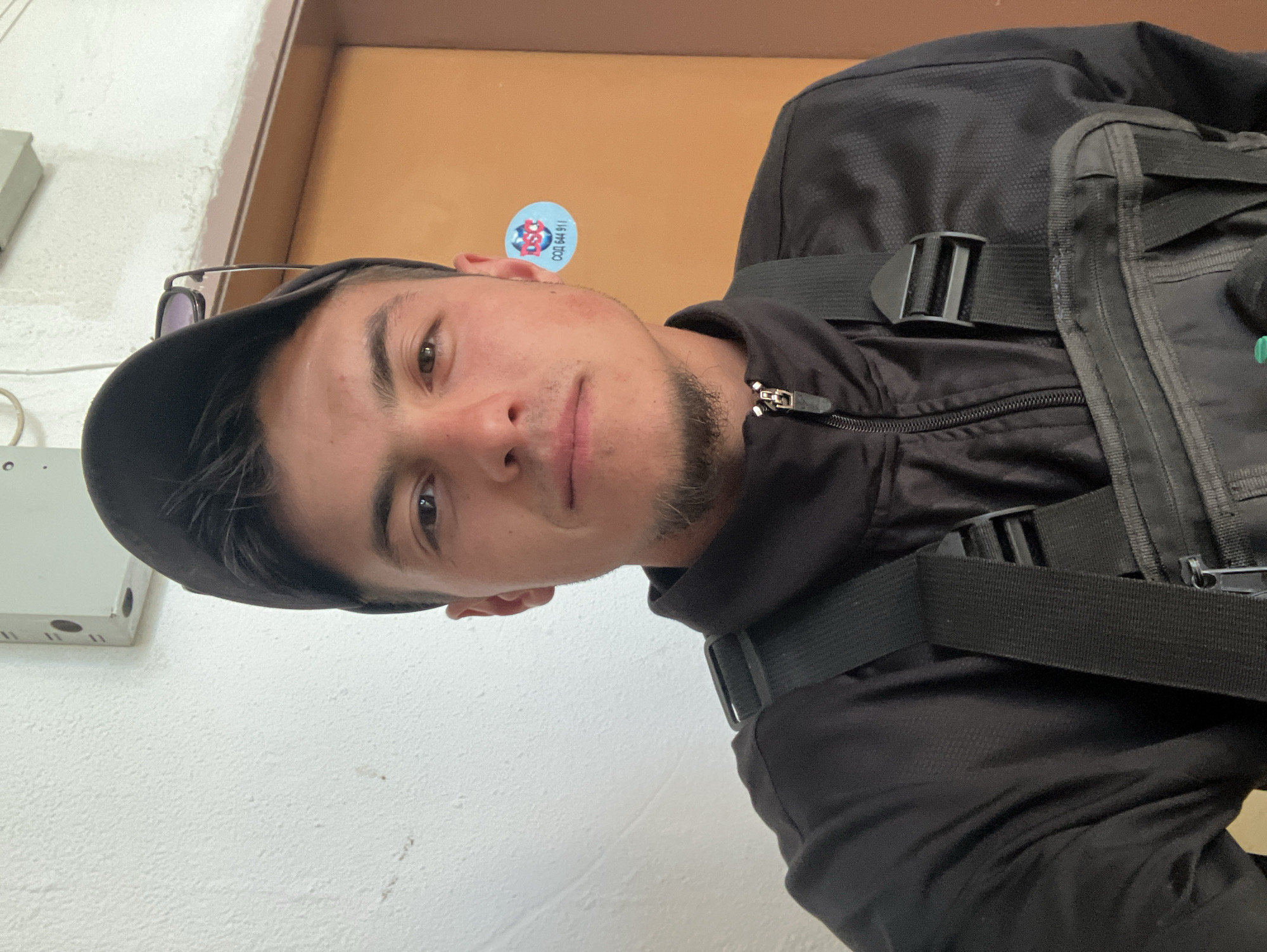 A selfie of a young man wearing a black cap, black jacket, and black backpack. The man is fair-skinned with brown hair, a trimmed beard, and dark eyes. He appears to be indoors, standing in front of a white wall and a light brown door. A small sticker with "СОД 644 911" (likely an emergency or security services number) and an emblem is affixed to the door. A white electrical box with wires is on the wall near the top left of the frame. Without additional information, the specific location in Plovdiv, Bulgaria, cannot be determined.