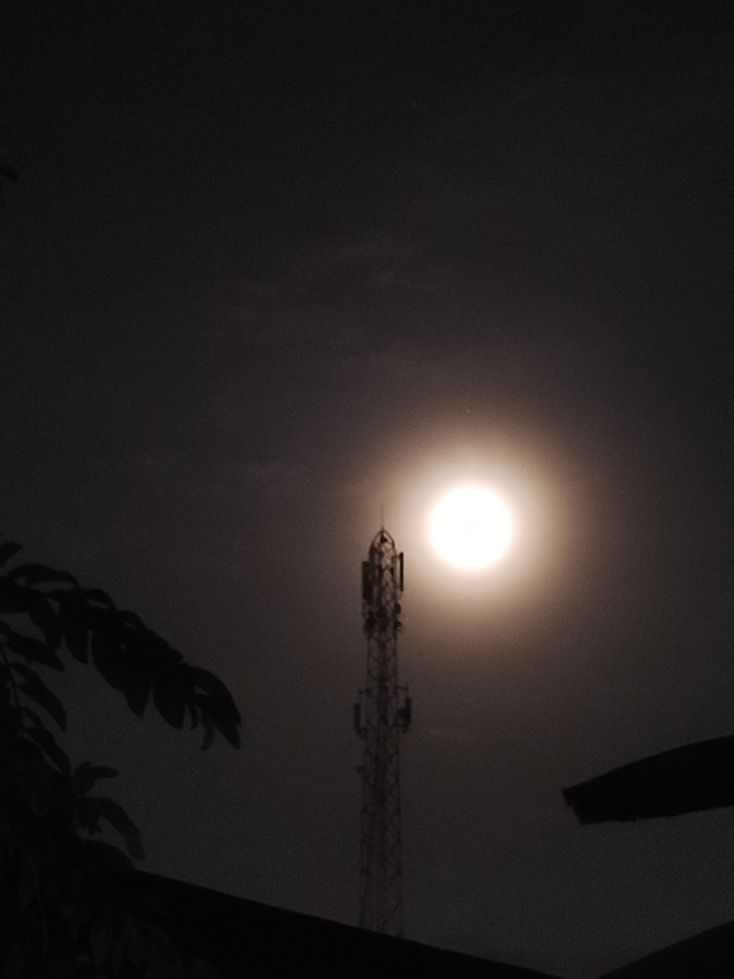 The image shows a night scene in Lhokseumawe, Indonesia. A cell tower is positioned centrally, silhouetted against a bright, full moon. To the left, dark foliage partially obscures the view. The lower portion of the frame is dominated by dark, indistinct shapes possibly representing rooftops or other structures. The sky is dark with some cloud cover. No people are visible.