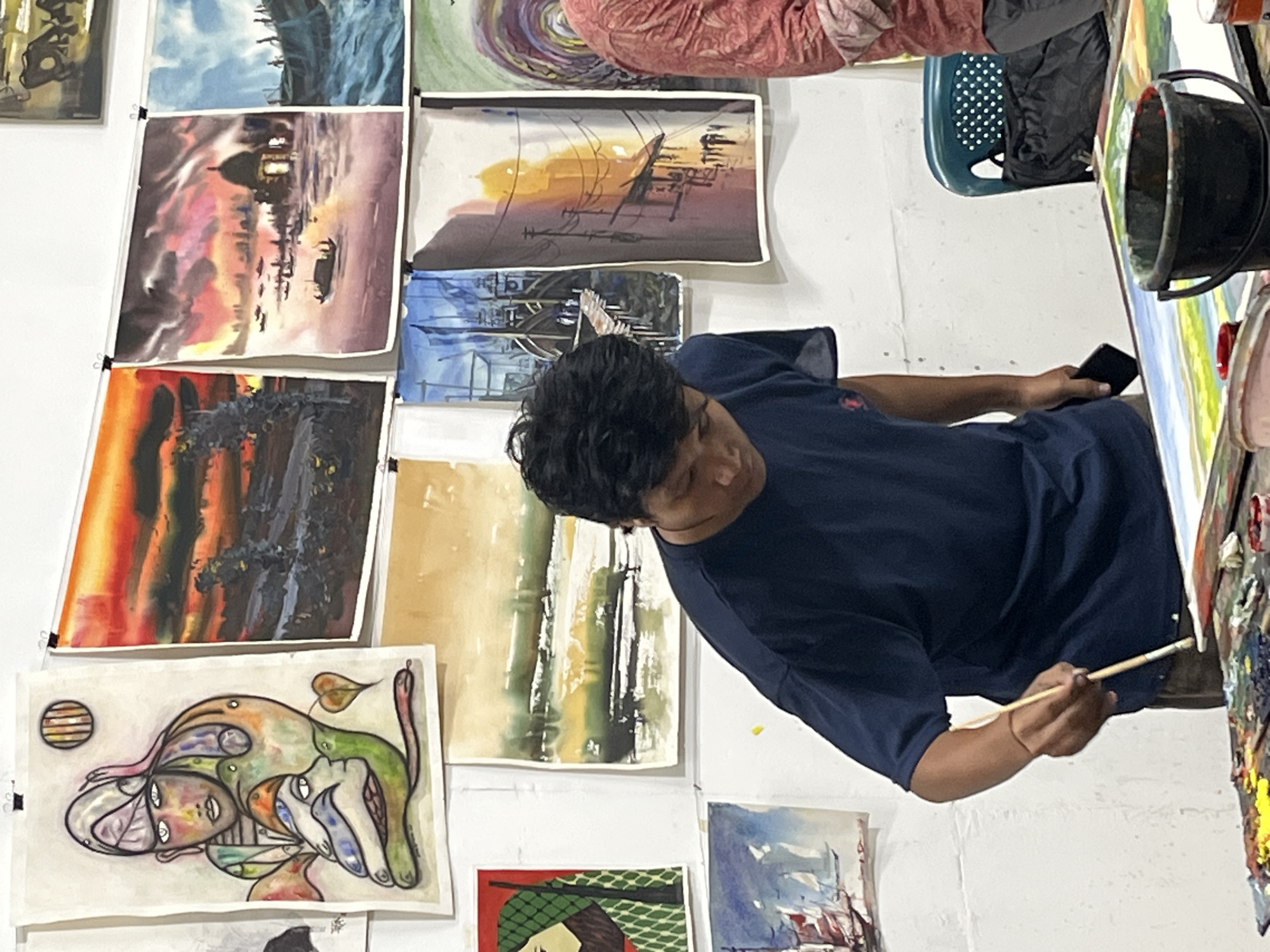A man in a navy blue T-shirt is seen painting in a room with numerous paintings displayed on the white wall. The paintings vary in style and subject matter, including landscapes, abstracts, and cityscapes. The man is holding a paintbrush in his right hand and a phone in his left. He is focusing on his painting, which is propped up on an easel to his right, and next to him is a black bucket. There is a blue plastic chair in the background. The location is presumed to be in Dhaka, Bangladesh, based on visual context.