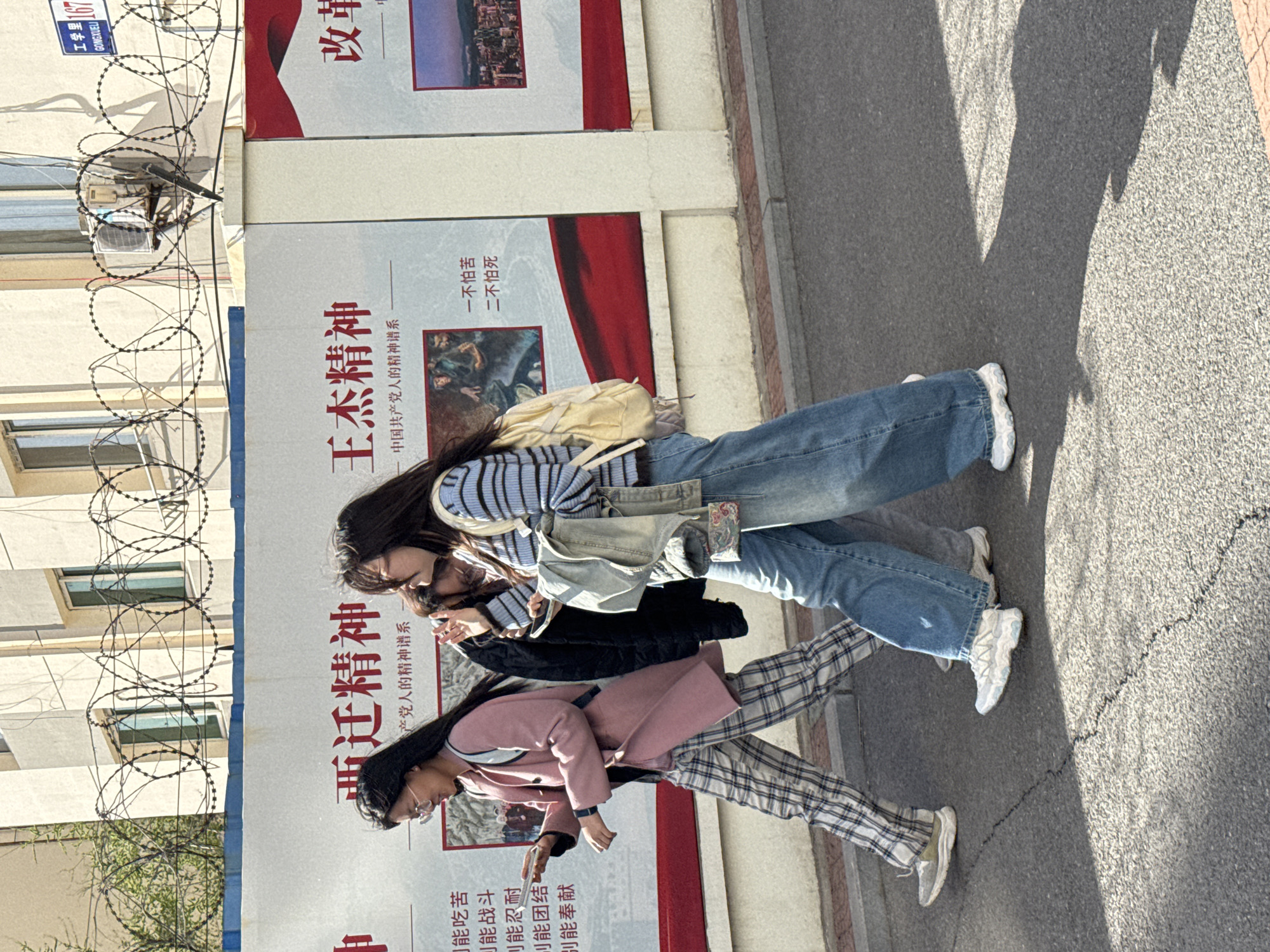 Three young women walk along a sidewalk next to a road in Zhongtun, China, under clear, sunny daylight, indicated by strong shadows. The women are casually dressed. The woman in the foreground wears a pink coat and plaid trousers, looking down at a mobile phone. Behind her, another woman in a dark jacket also appears to be focused on a device in her hands. The third woman, at the back, is dressed in a striped top, wide-leg blue jeans, and carries a light-colored backpack and a tote bag.
To their left, a wall is adorned with large propaganda-style banners featuring Chinese text and images. Barbed wire runs along the top of this wall. A blue sign on the wall reads "工学里 167 GONGXUELI".
Visible text on the banners includes phrases like "王杰精神" (Wang Jie Spirit), "中国共产党人的精神谱系" (Spiritual Genealogy of Chinese Communists), "一不怕苦 二不怕死" (First, not afraid of hardship; Second, not afraid of death), and "改革" (Reform). Another banner mentions "亚迁精神" and lists traits such as "能吃苦" (can endure hardship), "能战斗" (can fight), "可能忍耐" (perhaps can be patient), "可能团结" (perhaps can unite), and "别能奉献" (don't dedicate). The overall scene depicts urban life with political messaging prominent in the background.