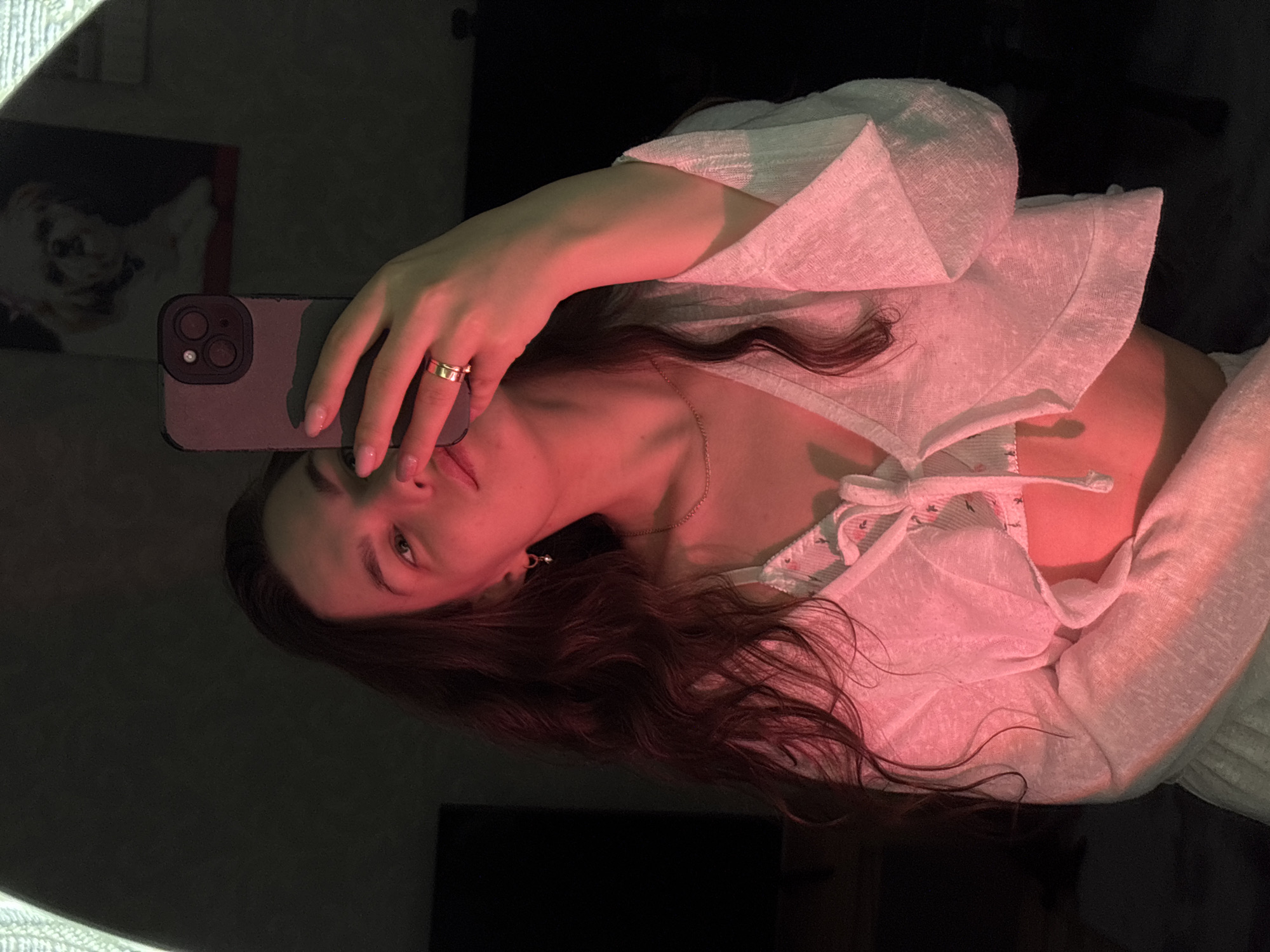 The image shows a young woman taking a selfie in what appears to be a bedroom. She is wearing a white, slightly sheer, cropped top over a patterned bralette and a white skirt. Her hair is long and dark, falling over her shoulders. She is wearing a gold ring on her right hand and a small hoop earring in her left ear. The lighting in the room is predominantly red, casting a strong hue across her face and clothing. In the background, a framed picture of a small white dog is visible on the wall. The overall impression is of a casual, personal moment captured at home. The location is identified as Chaikovsky, Russia.