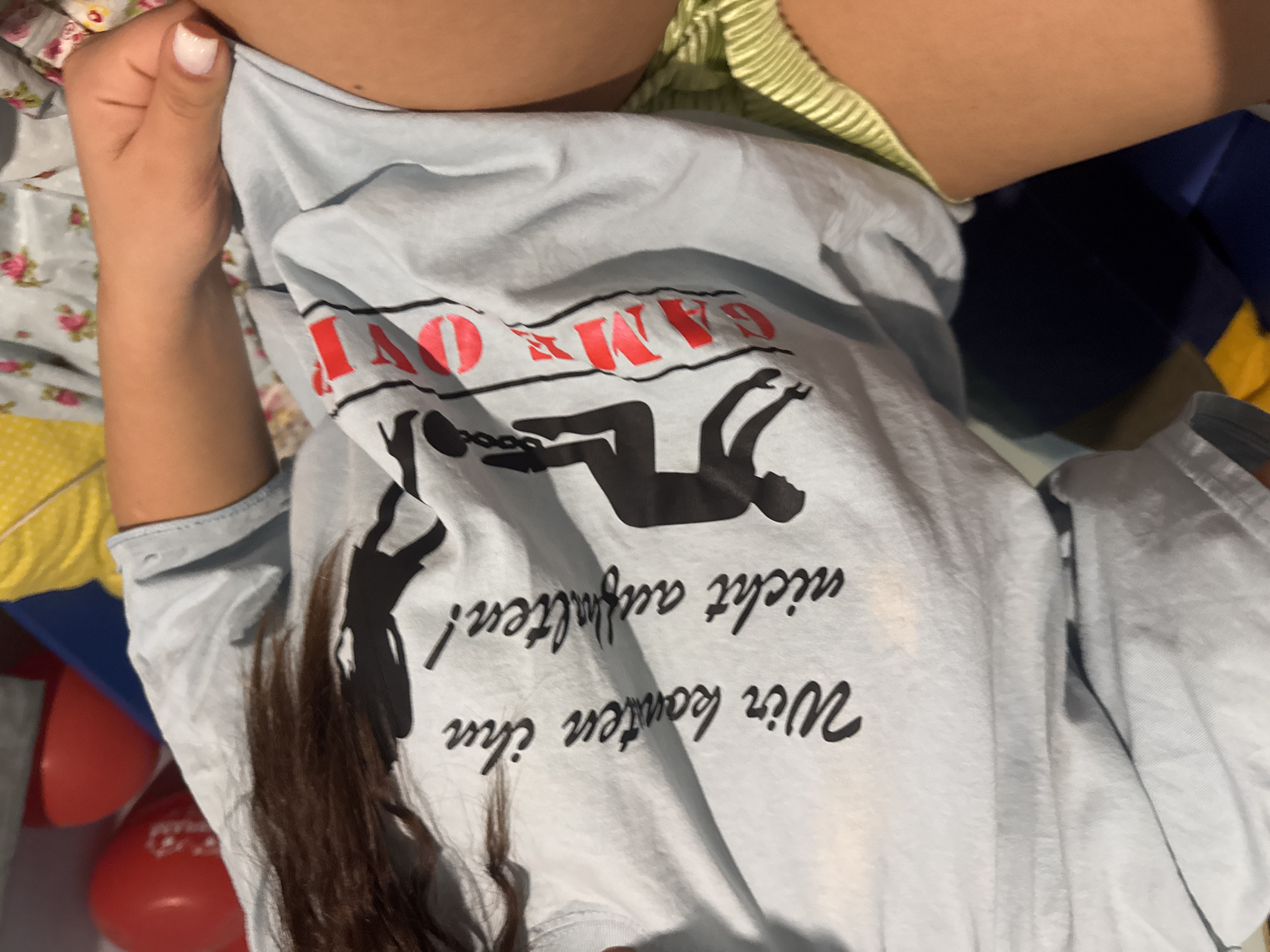 The image shows a person wearing a light blue t-shirt with a graphic of two figures chained to each other. Above the figures is the text "GAME OVER" in red. Below the figures is German text, "Wir konten ihn nicht aufhalten!". The person is lying down on what appears to be a bed with a floral pattern sheet and a yellow object in the background. There are also red objects in the background. The location is Unknown, Unknown.