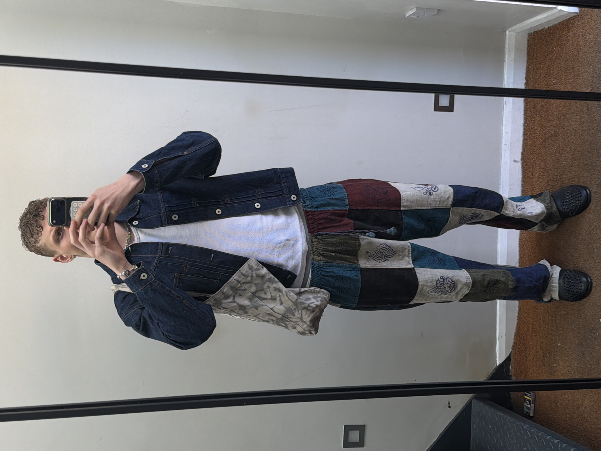A young person is captured in a mirror selfie indoors. The individual, likely male, has short curly brown hair and is seen holding a black smartphone to take the picture, with their face partially obscured. They are dressed in a dark blue denim jacket worn over a plain white t-shirt. A silver chain necklace and a bracelet are visible accessories. The person's left hand holds a grey patterned fabric tote bag or pouch.
The lower half of their attire consists of distinctive patchwork pants, composed of numerous square and rectangular fabric patches in various colors such as maroon, dark blue, light blue, grey, and black. Some of these patches feature intricate swirling or paisley-like patterns. The person is wearing black, textured, slip-on shoes.
The setting is a room with plain white walls, reflected in a large mirror with a dark frame. On the right side of the image, the floor is covered with a brown textured material, possibly carpet, alongside a dark grey structure that suggests a step or stairs. A white light fixture is visible on the ceiling, and a square vent or outlet is on the wall. The lighting appears bright and even. No visible text is present in the image.