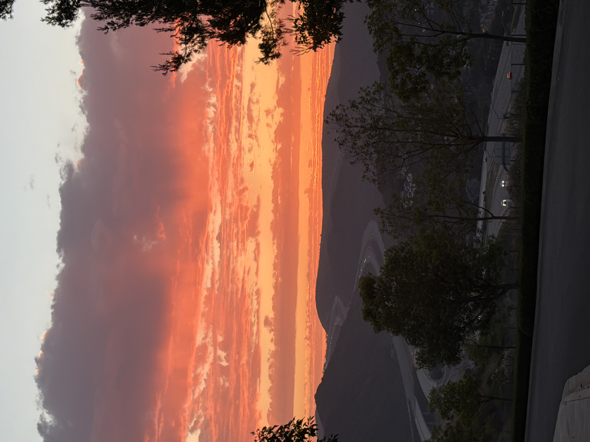 A vibrant sunset paints the sky with hues of orange and red, casting a warm glow over the rolling hills of Aliso Viejo, California. The clouds are streaked with fiery colors, creating a dramatic contrast against the darkening landscape below. A winding highway snakes through the valley, its lanes illuminated by the headlights of a few cars, suggesting the transition from day to night. Trees sparsely dot the hillsides, their dark silhouettes standing out against the illuminated sky. In the foreground, a paved road and a low hedge suggest a viewpoint or park area overlooking the scene. The overall impression is one of natural beauty and the quiet hum of urban life at dusk.