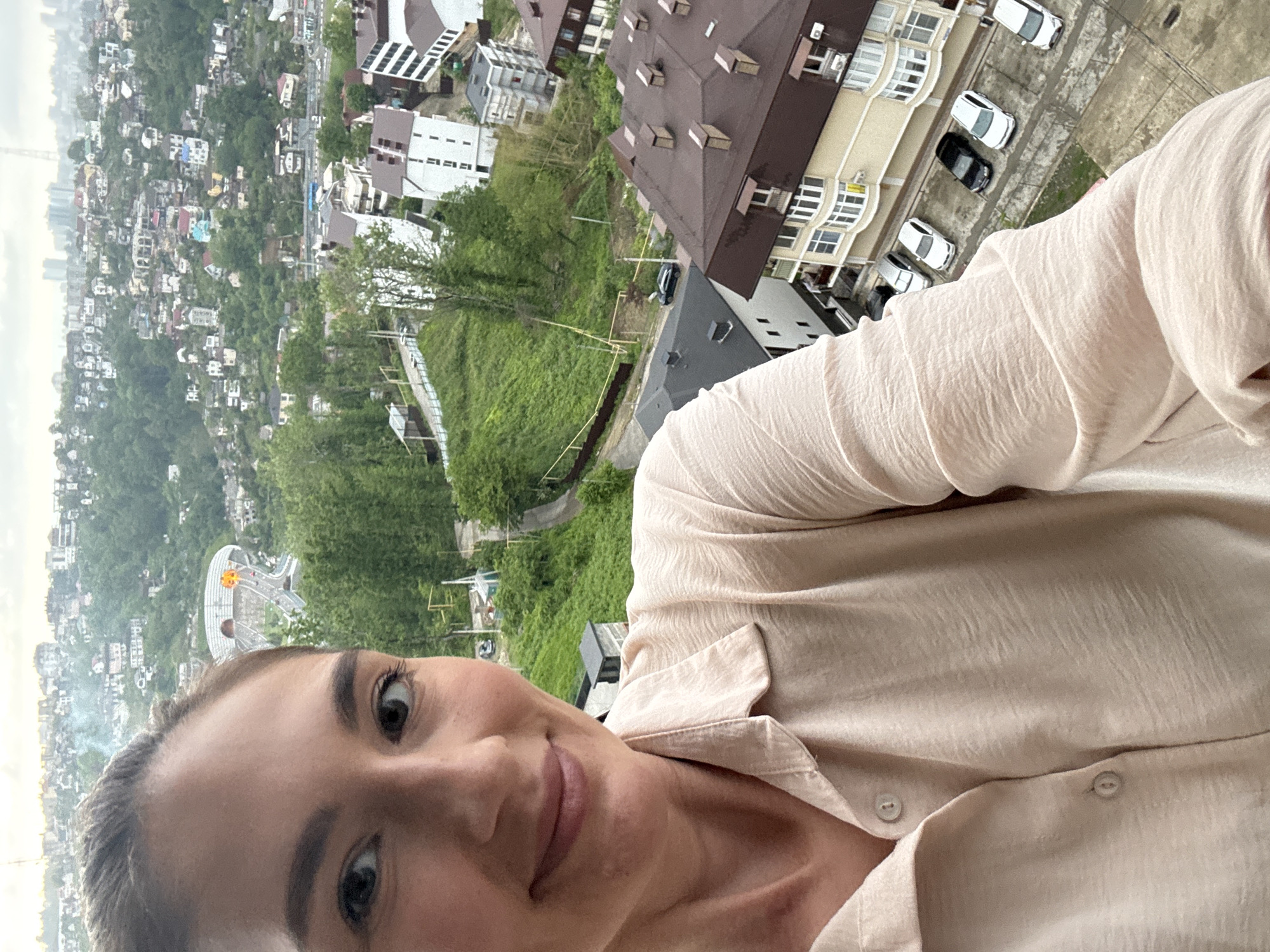 A selfie taken by a person with brown hair and fair skin, wearing a light tan, button-down shirt. The subject is smiling slightly. The background shows an elevated view of the city of Sochi, Russia, with numerous buildings, trees, and green spaces visible. A Ferris wheel can be seen in the distance on the left. Several cars are parked near buildings with brown roofs in the mid-ground. The sky is overcast.