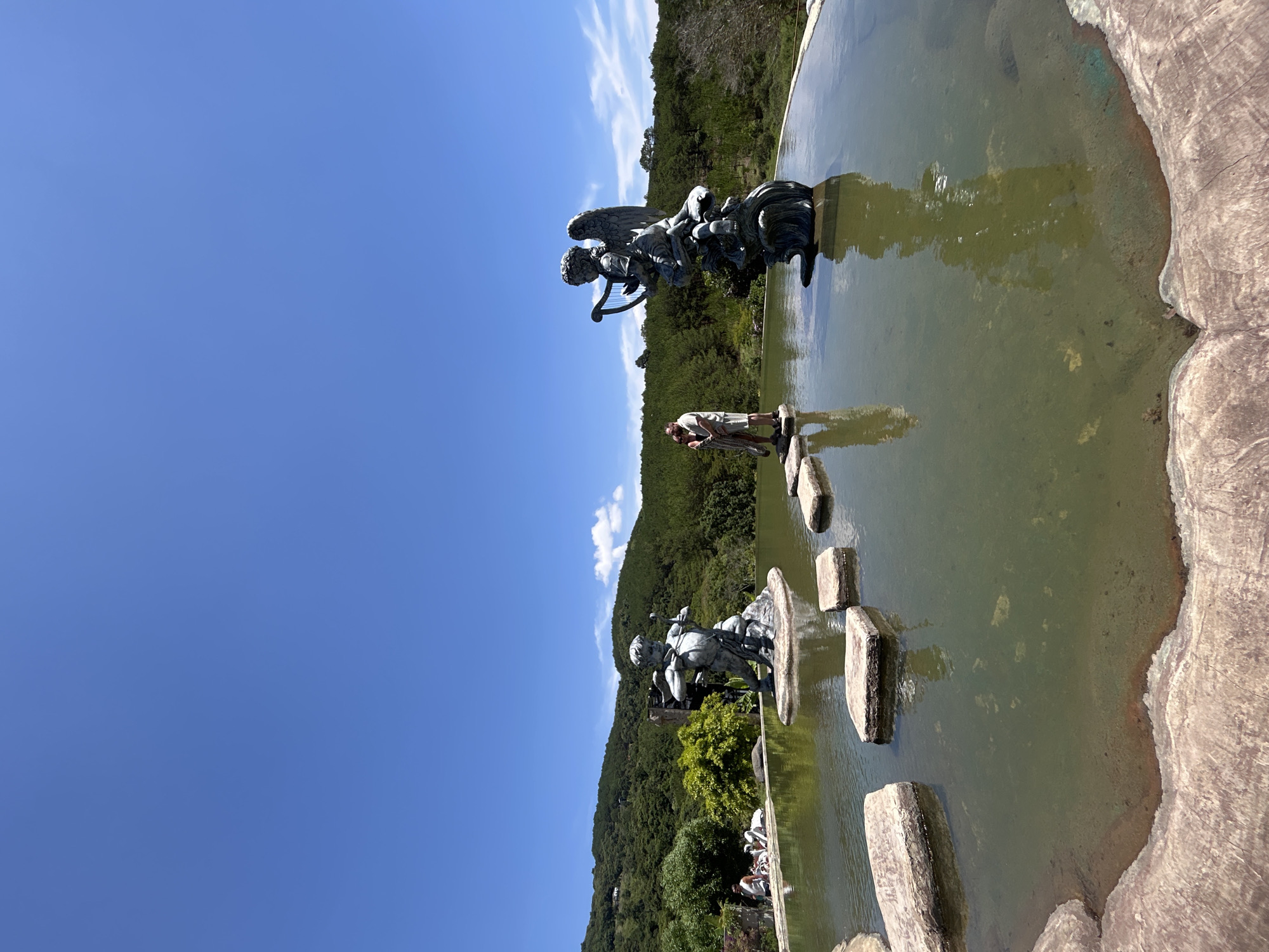 The image captures a serene outdoor scene, depicting a sculpted garden or park area, likely in Da Lat, Vietnam, under a bright, clear sky.
A wide, shallow pool of greenish-brown water fills the foreground and midground, suggesting the presence of sediment or algae at the bottom. A path of large, irregularly shaped stone stepping stones traverses the water, leading into the distance.
Within or beside the water, several dark-colored statues are prominent. On the left side of the frame (when upright), a statue depicts a figure, possibly a cherub, riding on an animal or chariot and holding a staff or weapon. Further to the right, another ornate statue portrays an angelic figure with large wings, appearing to play a harp while kneeling or seated.
One person is visible standing on a stepping stone in the water, looking downwards, potentially observing the water or adjusting something. In the background, at the base of a verdant, forested hill, several other indistinct figures can be seen, indicating the presence of more visitors in this public setting.
The backdrop is a lush, green hillside covered with dense foliage, hinting at a natural or extensively landscaped environment. Above, the sky is a striking clear blue with only a few wispy white clouds. The strong, natural lighting suggests it is midday or early afternoon, with sunny weather. The overall ambiance is tranquil and inviting for sightseeing. No discernible text is visible within the image.