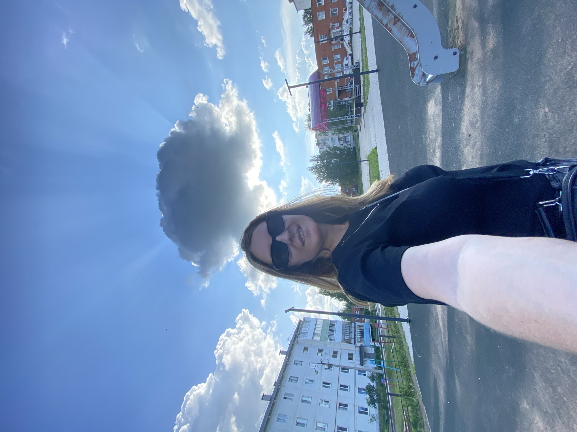 A woman wearing sunglasses and a black t-shirt is taking a selfie outdoors. She has long, blonde hair and is smiling. Behind her, a cloudy sky with sun rays is visible. In the background, there are residential buildings and a playground with a slide. The scene appears to be in the city of Noviy, Russia, based on the architecture and the playground setting. The woman's arm is extended, holding the camera, and she is positioned in the foreground. The overall atmosphere is casual and bright.