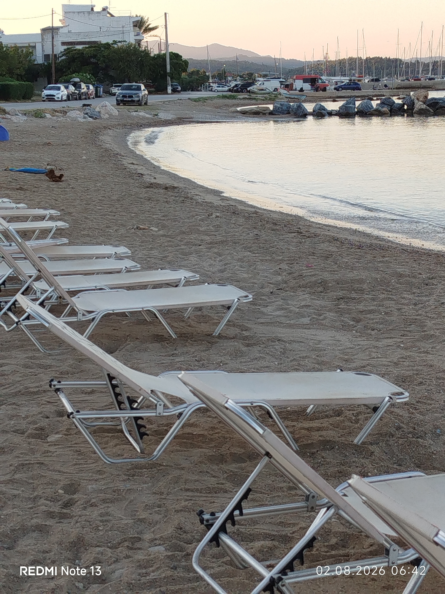 The image shows a beach in Sithonia Municipal Unit, Greece, with multiple empty lounge chairs lined up on the sand. The sea is calm and reflects the sky, with a rocky breakwater in the middle ground. In the background, there are parked cars, trees, and buildings, and beyond that, a marina with numerous sailboats. The sky is a pale orange, suggesting either sunrise or sunset. The overall scene is peaceful and unpopulated, with the focus on the arrangement of the lounge chairs and the natural environment.