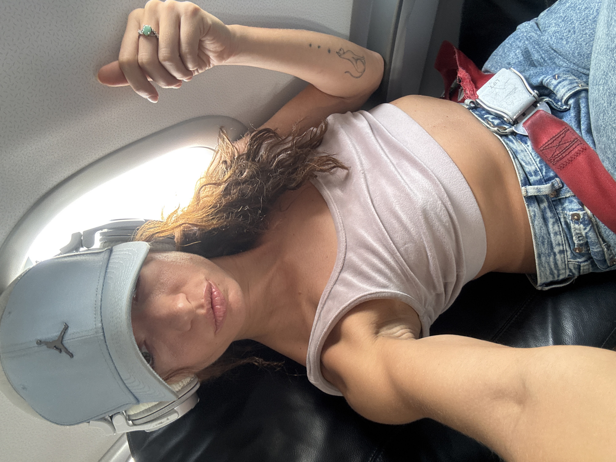 A person, likely female, is lying in what appears to be an airplane seat, wearing a light pink tank top, blue jeans, a blue baseball cap with a Jordan logo, and headphones. A red airplane seatbelt is fastened across the person's waist. A ring is visible on the person's left hand. A tattoo is present on the person's left forearm. The scene suggests the interior of an airplane. There is no evidence to pinpoint a specific location in Nong Prue Subdistrict, Thailand.