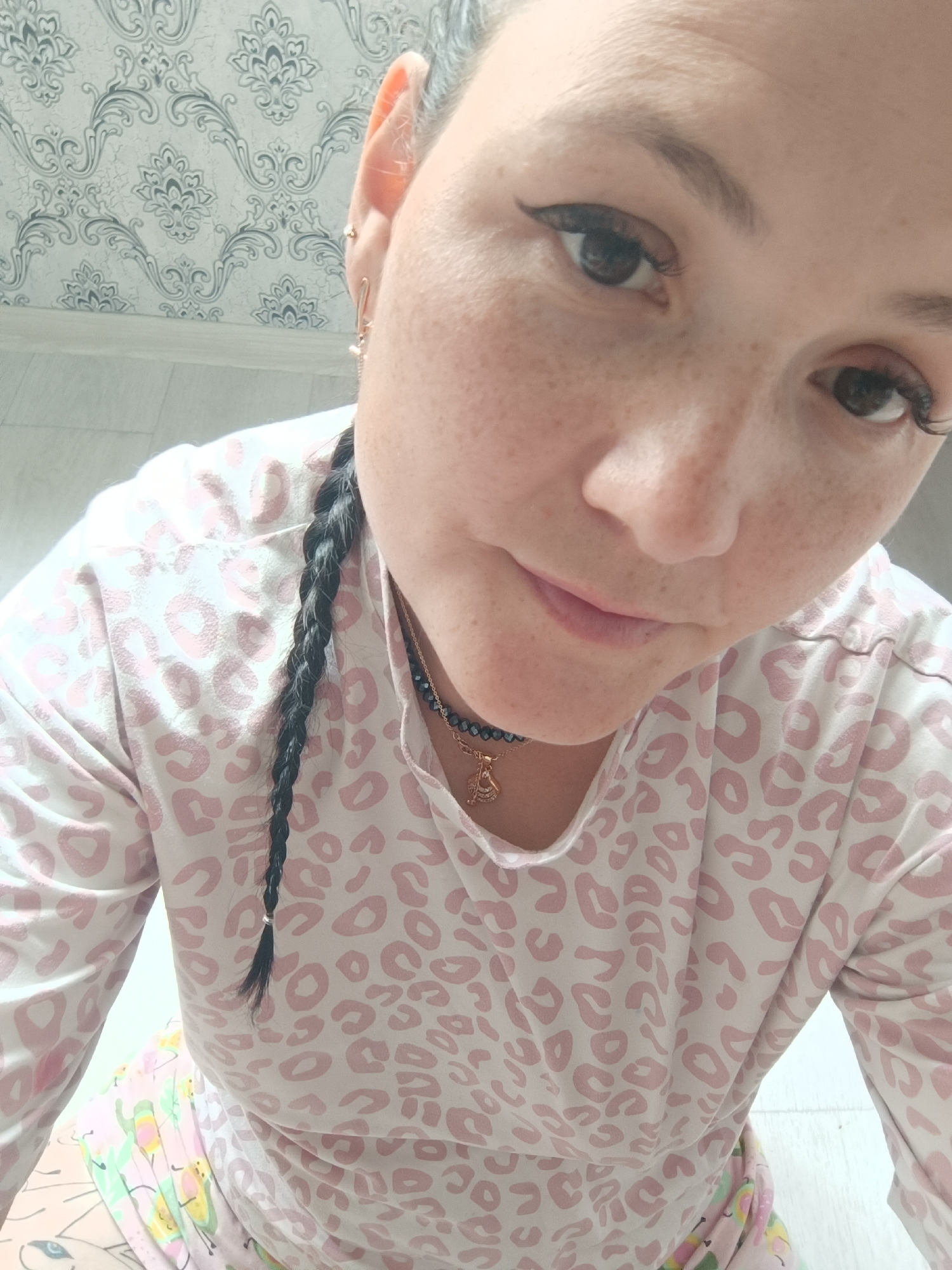 A close-up, overhead shot shows a woman with fair skin and freckles, wearing a pink and white leopard-print long-sleeved shirt. She has dark hair in a braid and is wearing a necklace with a gold pendant. The background appears to be a wall with floral wallpaper and a white floor, suggesting an indoor setting, possibly a home. The image lacks specific details to pinpoint a location in Chaikovsky, Russia.
