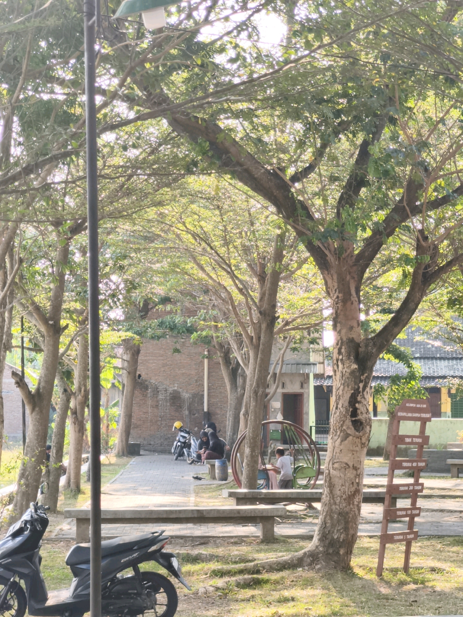 The image shows an outdoor scene in Desa Dibal, Indonesia, likely a public park or communal space. In the foreground, two dark-colored scooters are parked on a grassy area. A paved walkway extends into the background, lined by trees and brick buildings. Several people are seated on benches and near a playground structure, with two individuals on a swing set. A signpost with text in Indonesian stands to the right of a large tree. The overall impression is a peaceful, sunlit environment.